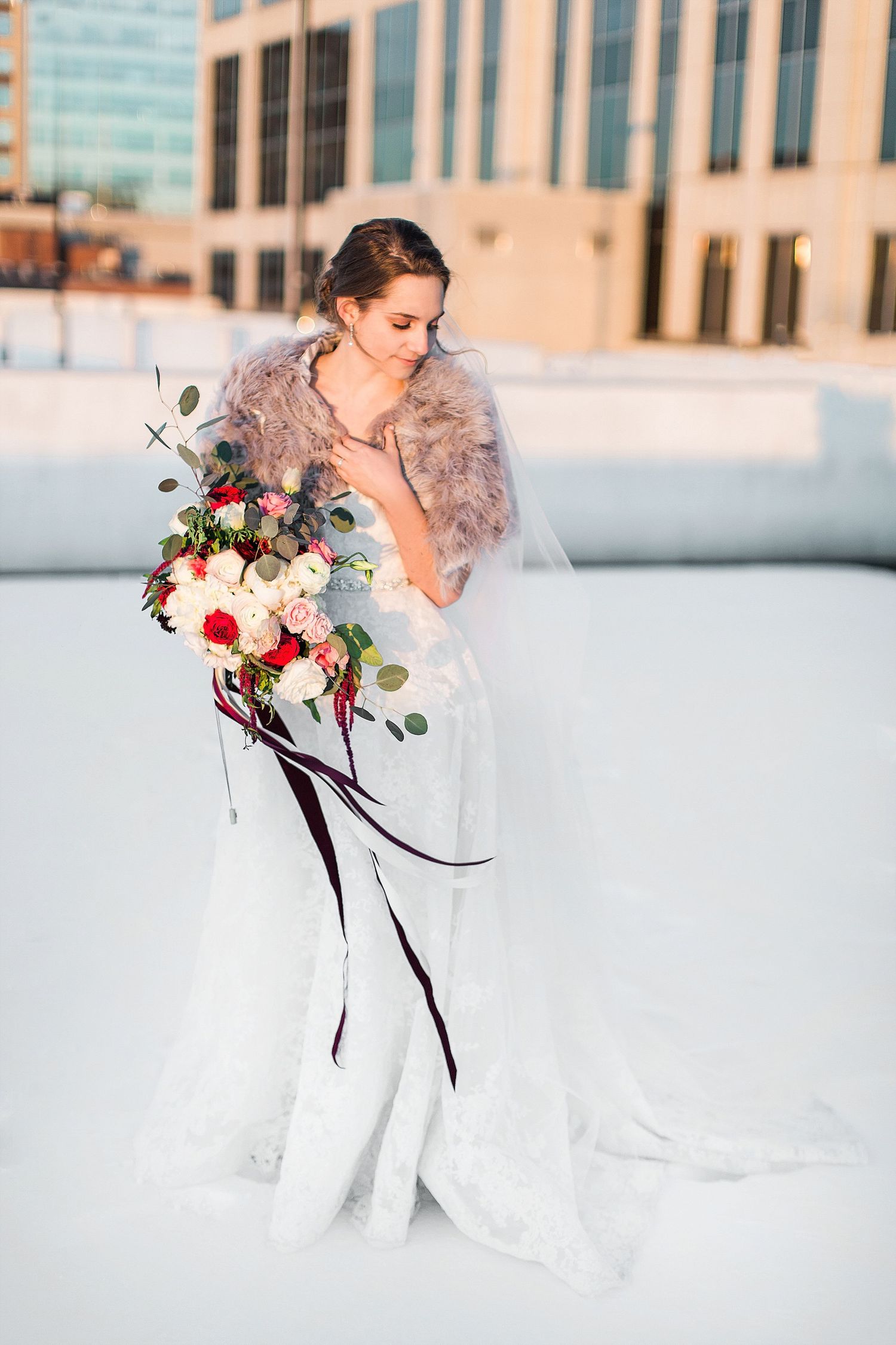 Bride in white dress with fur shawl holds cascading bouquet against snowy backdrop.