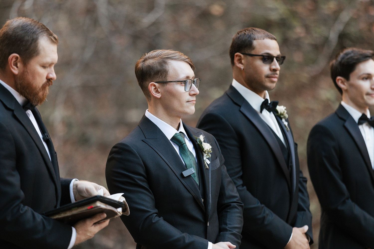 A group of groomsmen in black suits and sunglasses stand together during an outdoor wedding ceremony.