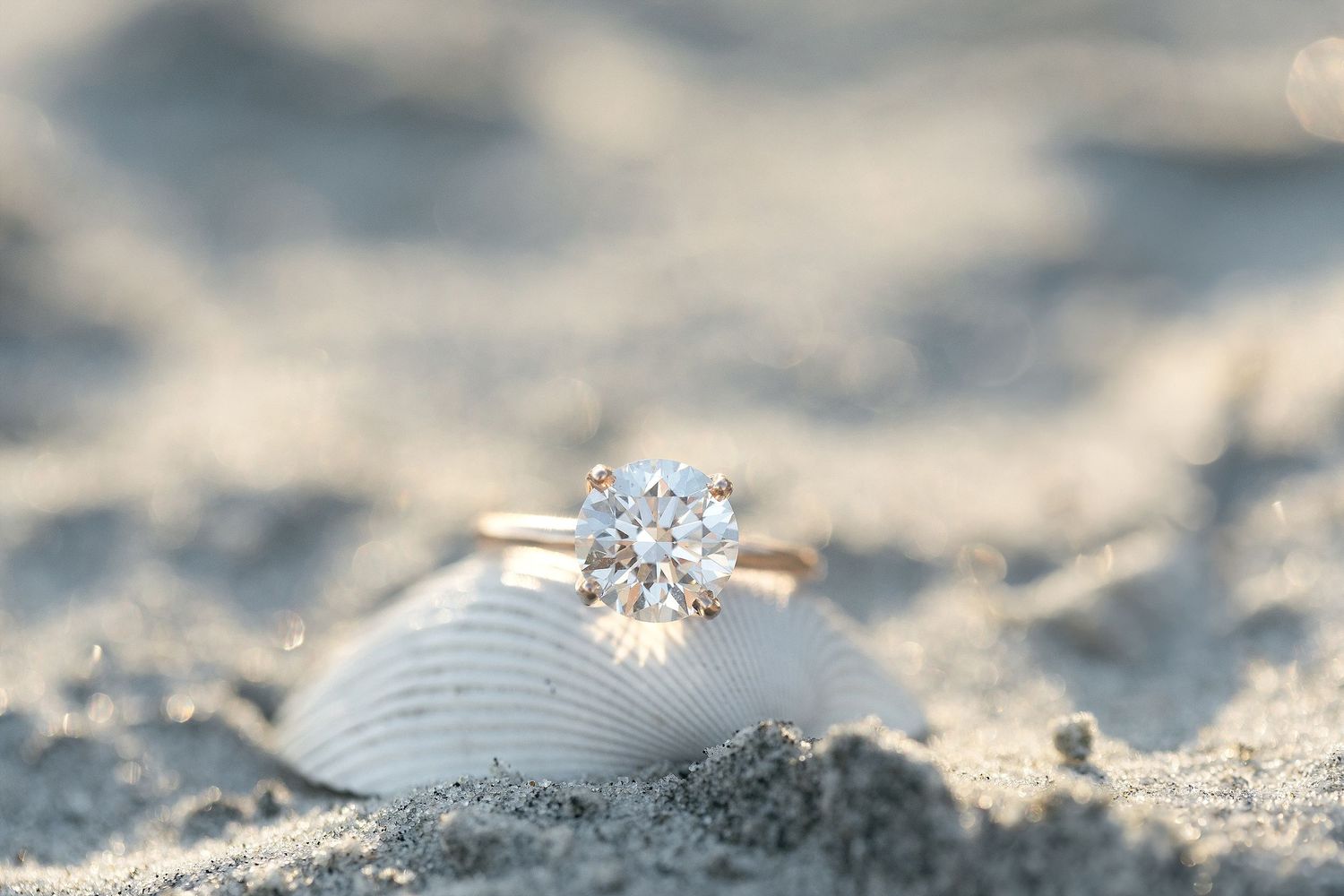 A sparkling diamond solitaire engagement ring rests on a white seashell in the sand with soft bokeh background.