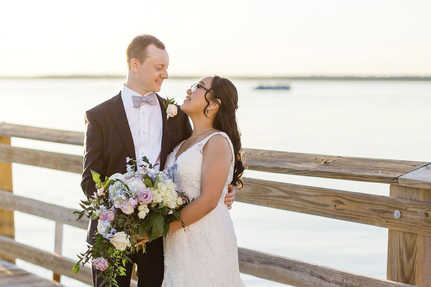 A couple shares an intimate moment on a waterfront pier with a beautiful floral bouquet.