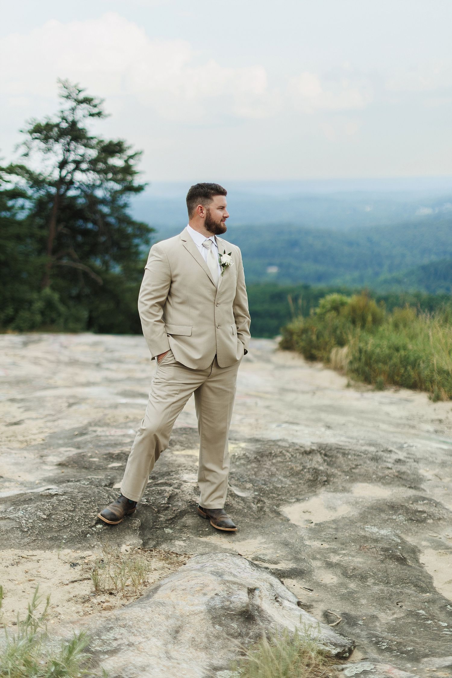 A groom in tan suit poses on rocky mountain overlook with forest valley below.