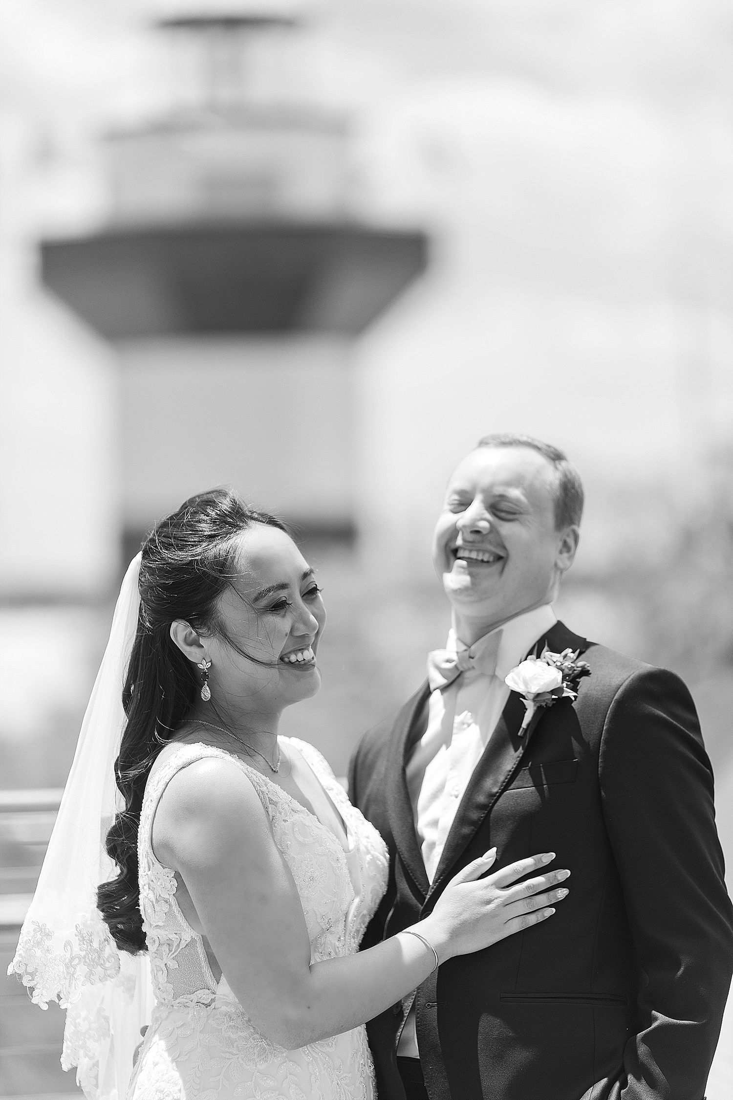 Black and white portrait of newlyweds laughing together with lighthouse silhouette in background.
