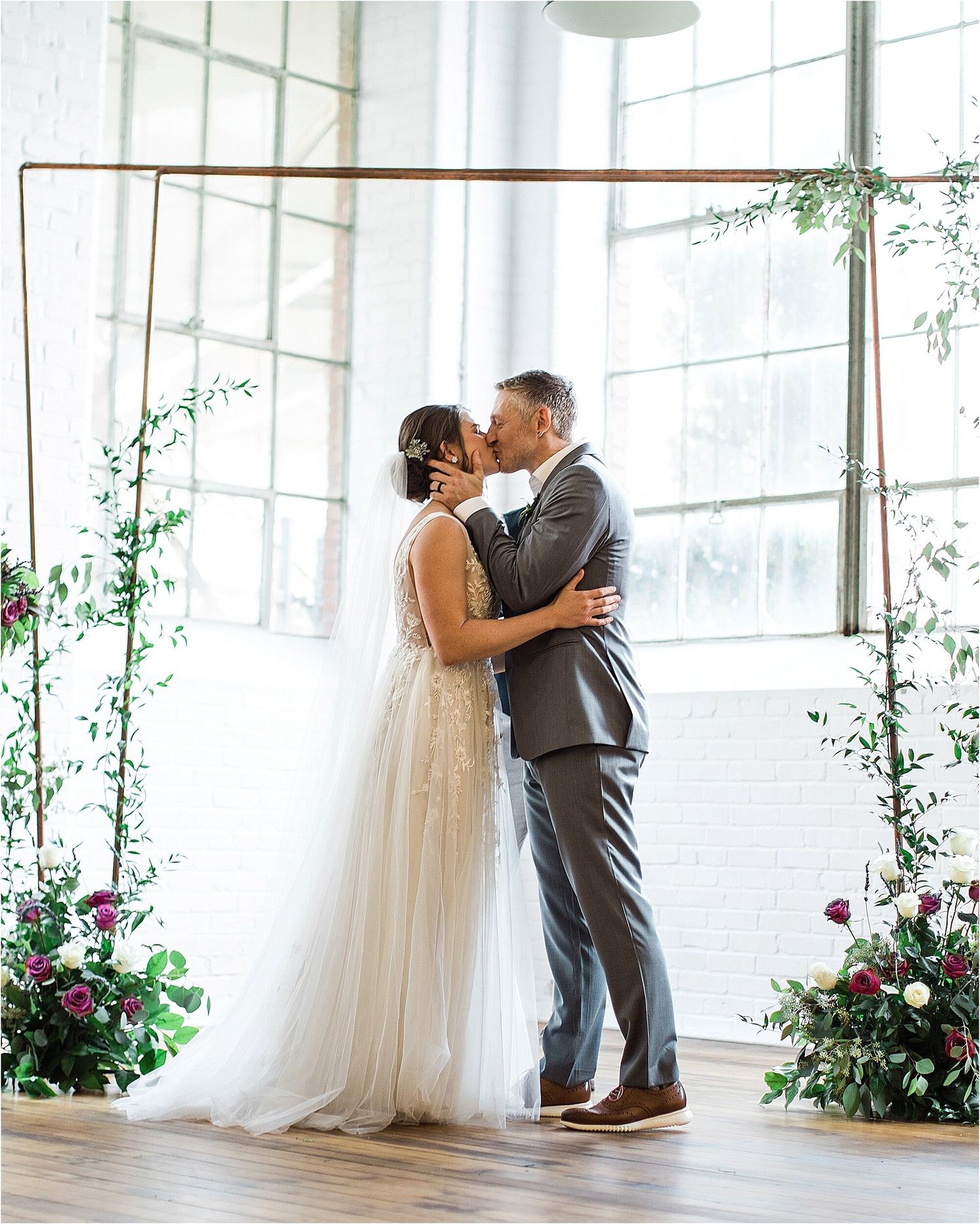 A romantic first kiss moment framed by floral arrangements and greenery at a wedding ceremony.