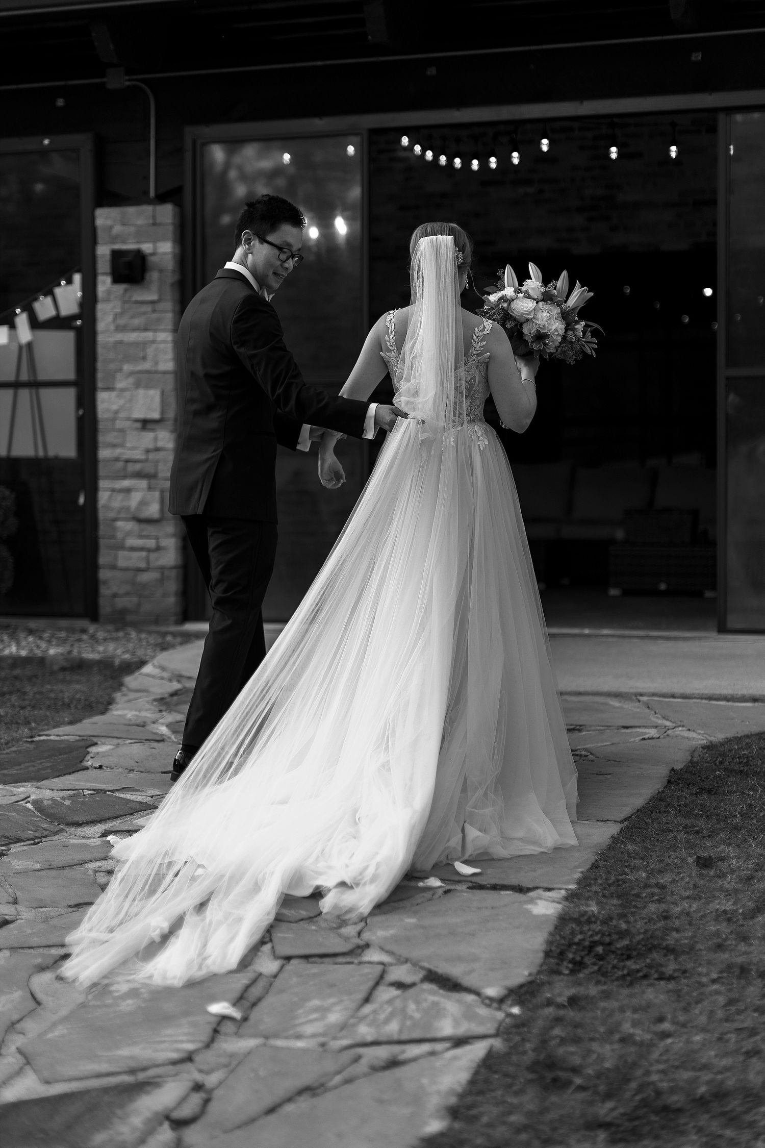 Black and white wedding photo showing two people on a stone patio with a long flowing bridal veil and gown.