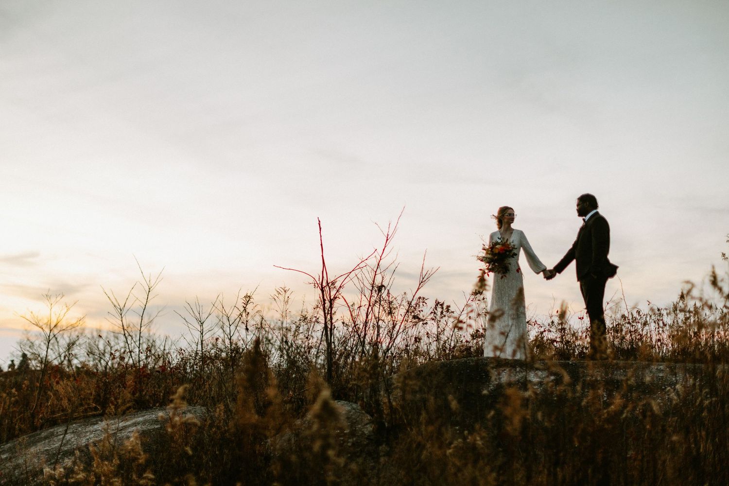 A couple stands silhouetted against a sunset sky on a hilltop surrounded by autumn foliage.