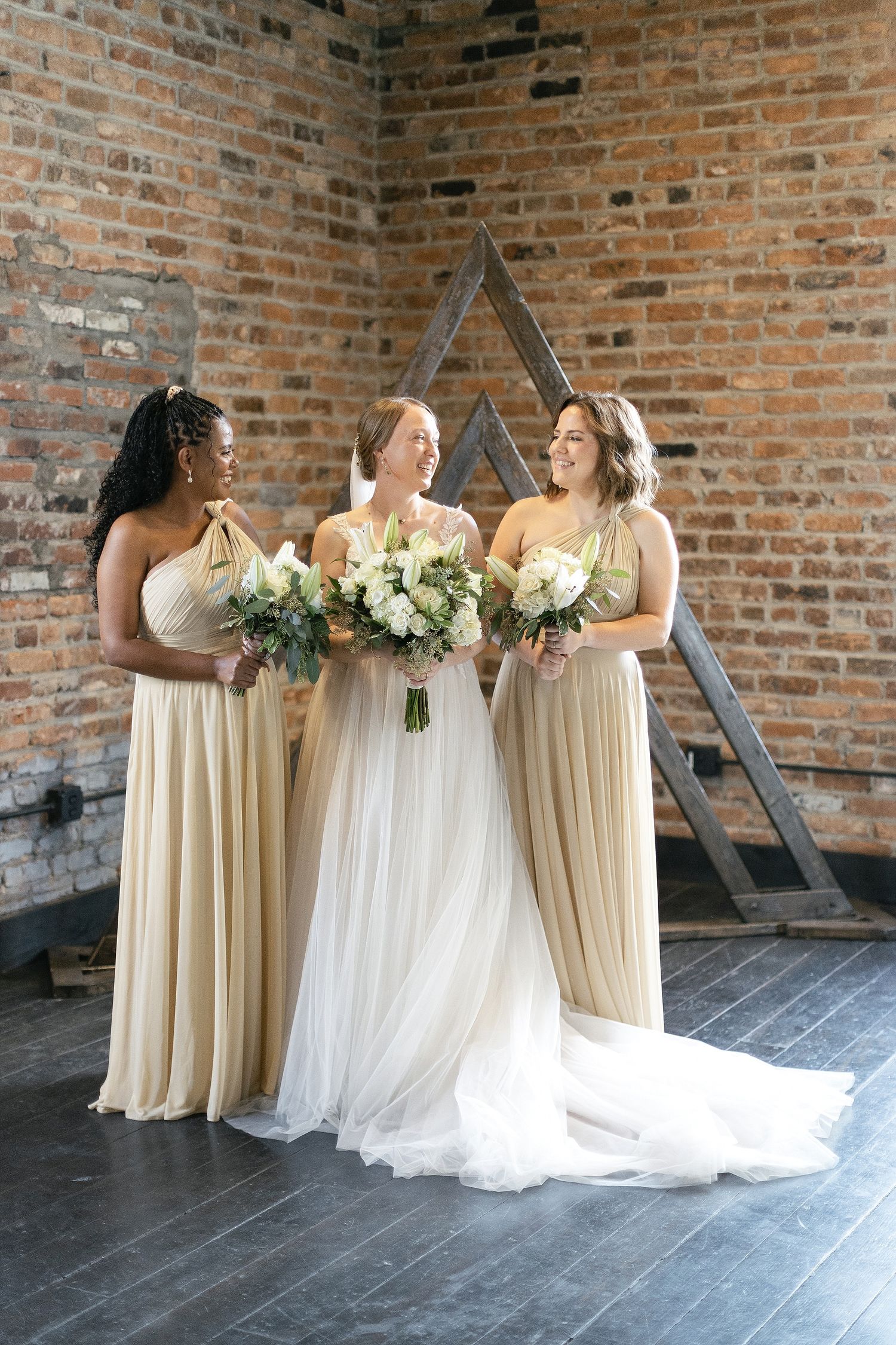 Three wedding attendants in cream-colored dresses pose together against a brick wall with elegant white bouquets.