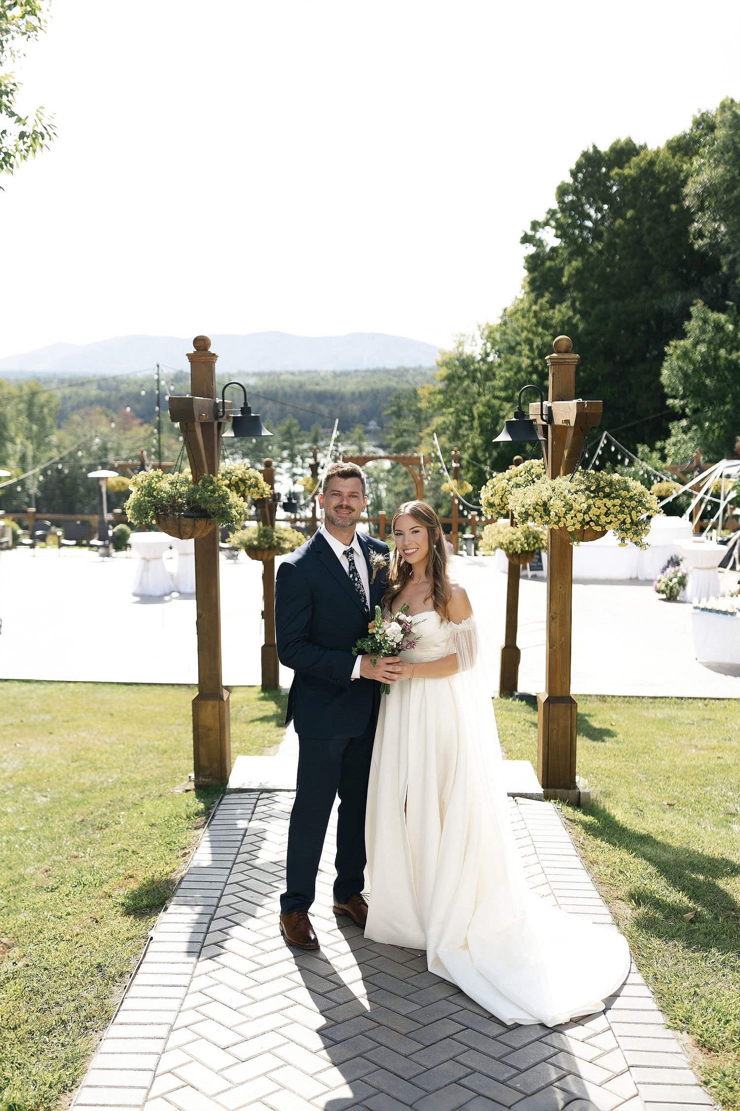 A wedding couple stands under a wooden arch decorated with flowers on a sunny day with mountain views.