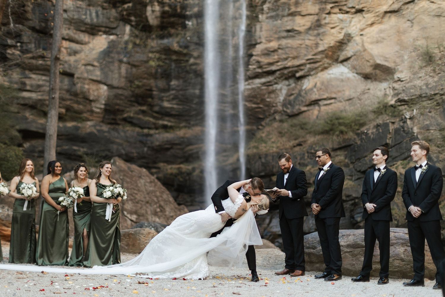 Wedding party celebrates with dramatic dip kiss in front of majestic waterfall against rocky cliff backdrop.