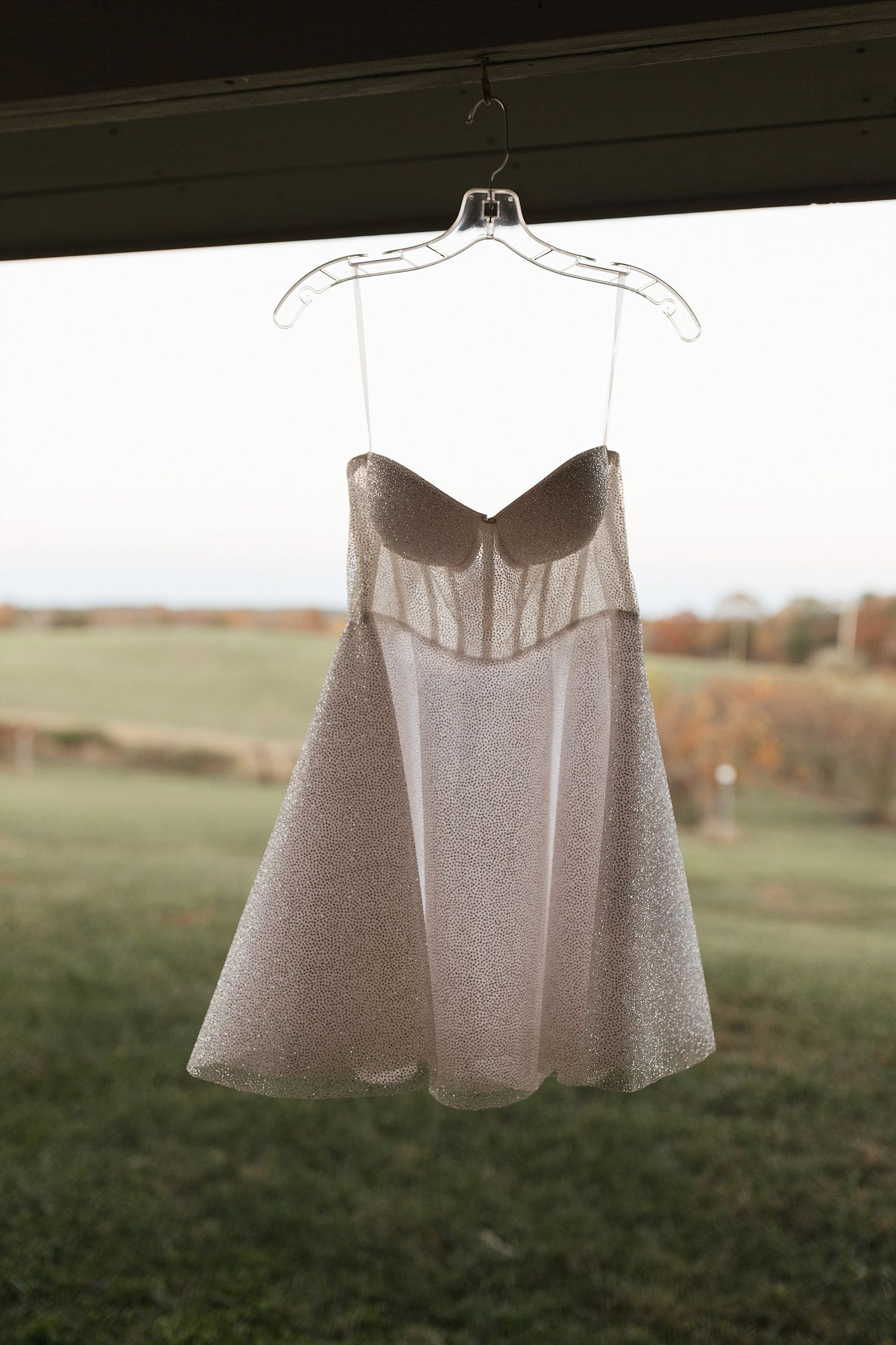 A delicate white wedding dress suspended from a hanger against a pastoral countryside background at sunset.