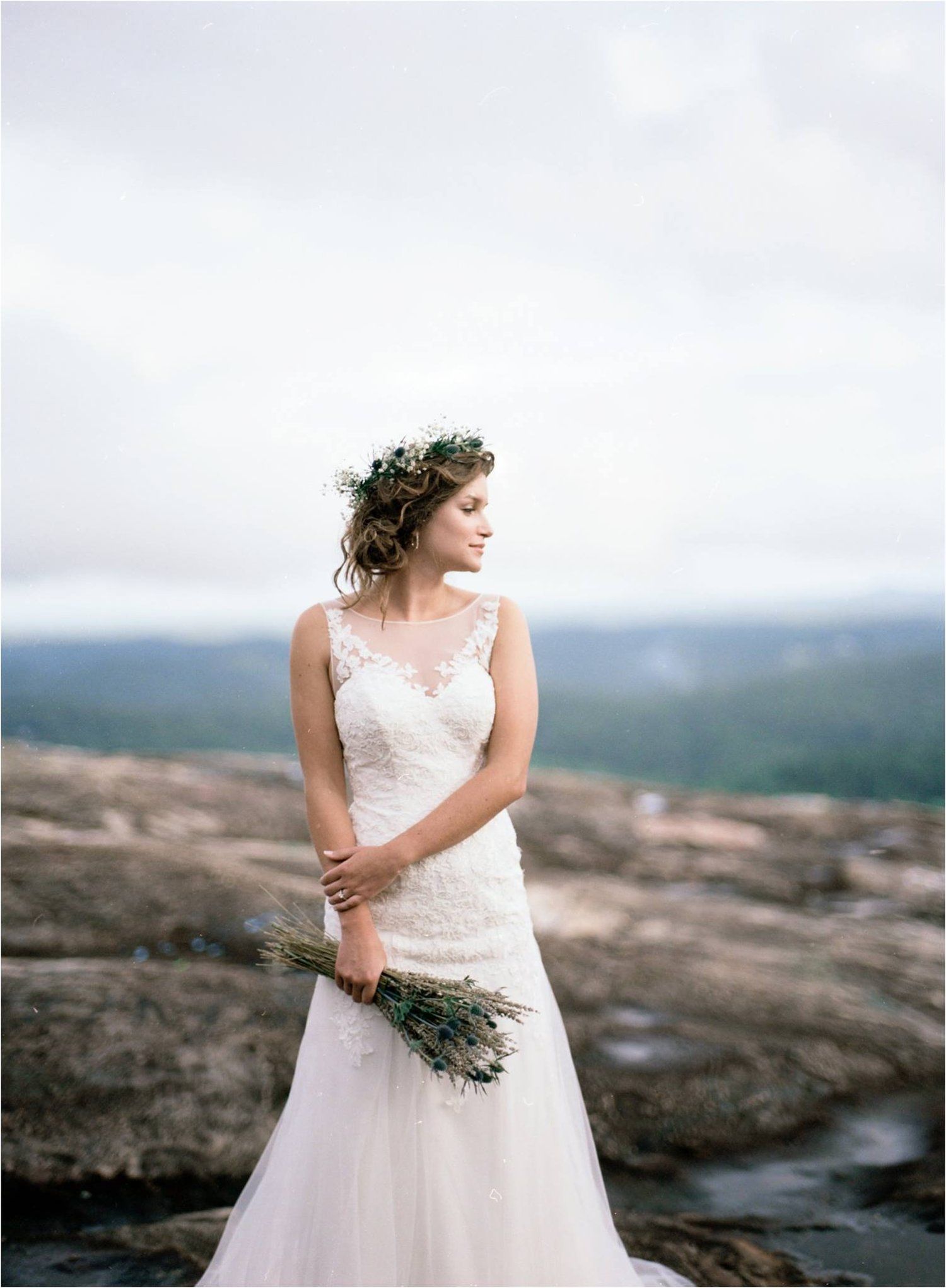 Bride in lace gown with floral crown poses on rocky overlook with mountain views.