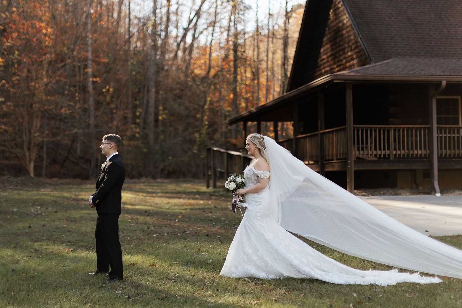 A rustic fall wedding scene in front of a wooden cabin with a long flowing white veil catching the autumn breeze.