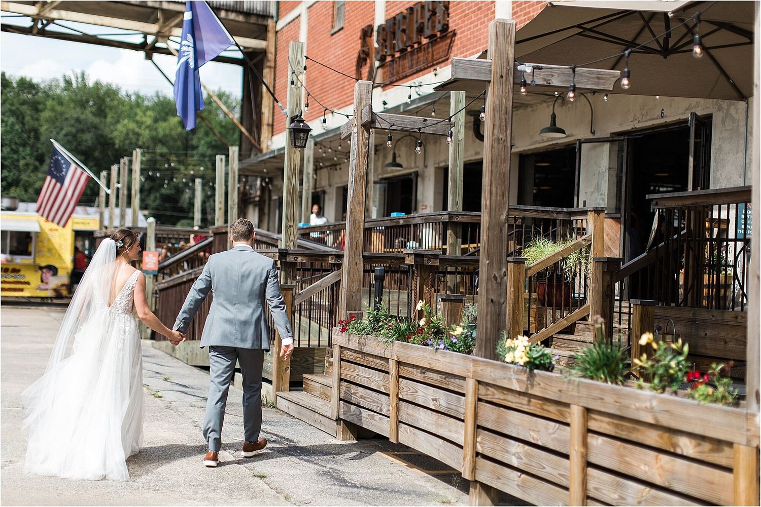 Newlyweds walk hand in hand past outdoor restaurant patios with American flags displayed.