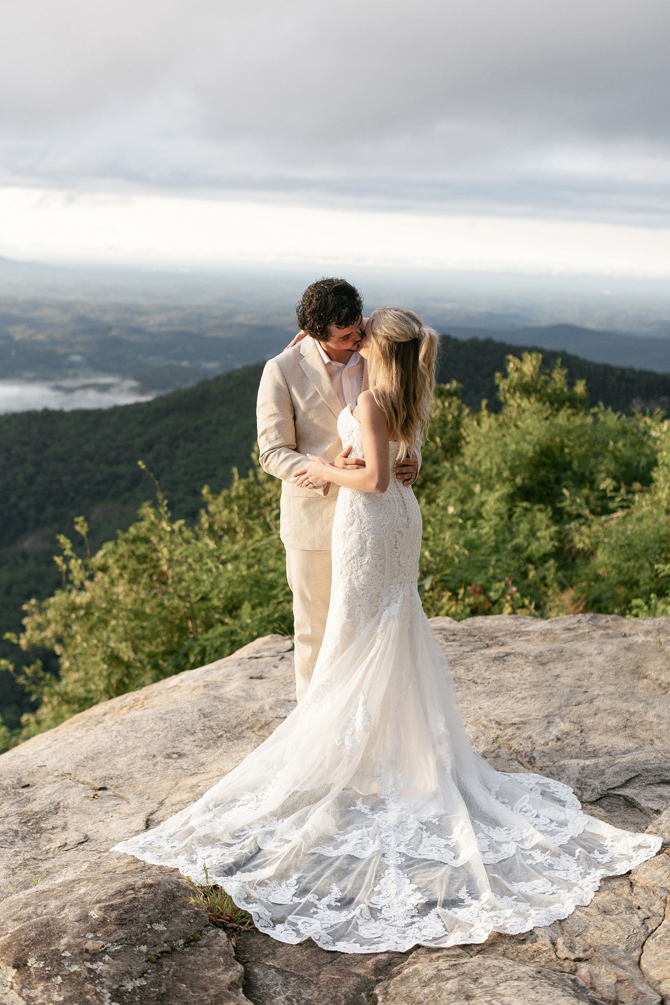 A romantic wedding couple shares intimate moments on a scenic mountain overlook with cloudy skies and lush greenery below.