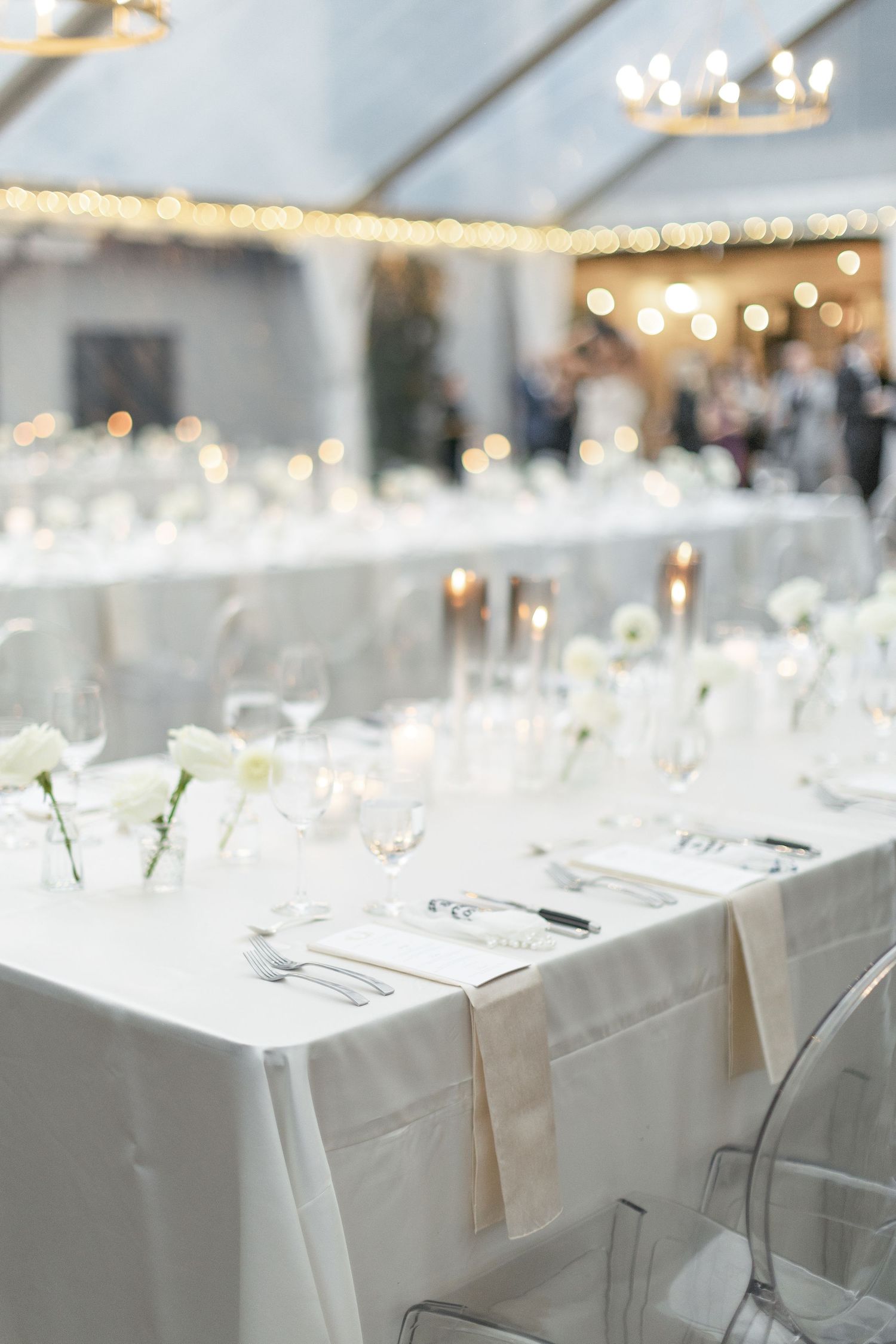 Elegant wedding reception table setting with white linens, clear chairs and soft bokeh lighting in the background.