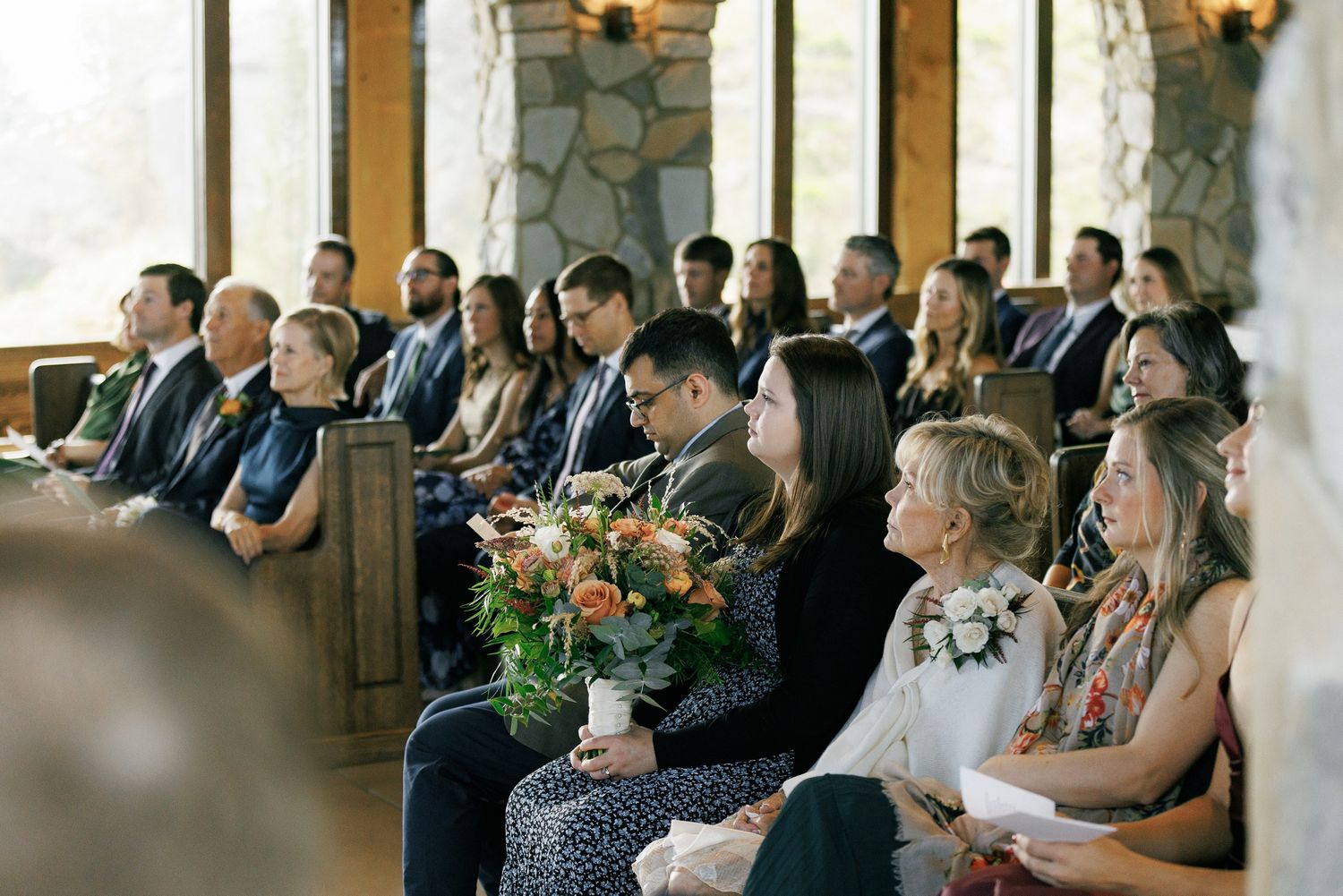 Wedding guests seated in rows at an indoor ceremony venue with stone walls and warm lighting.