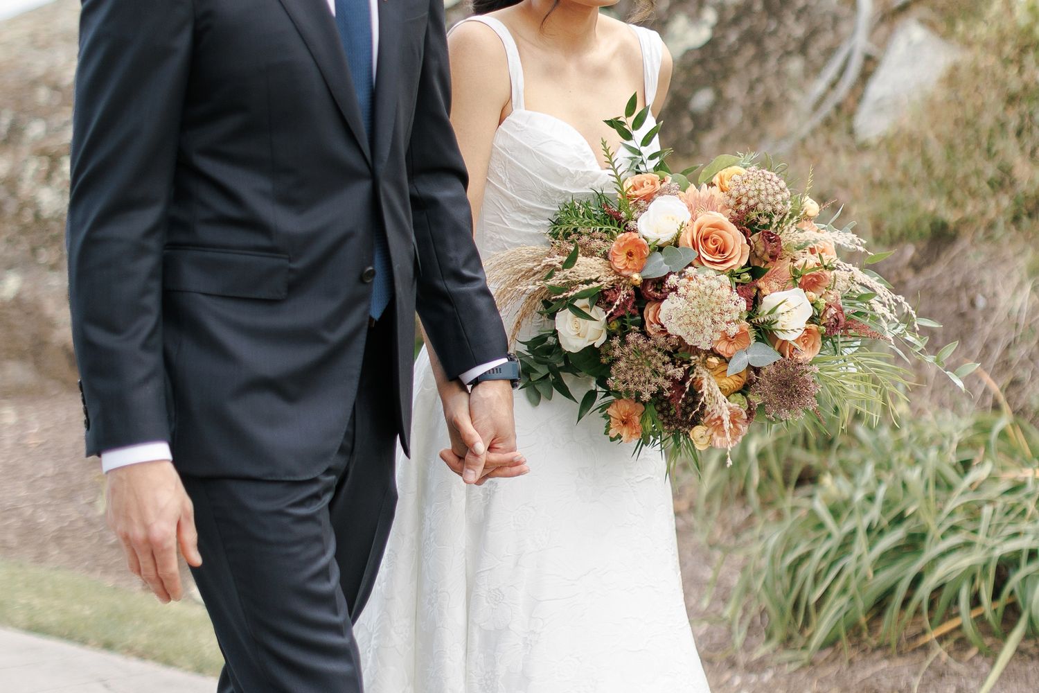 A wedding couple holds hands outdoors while the bride carries a lush peach and cream floral bouquet with greenery.