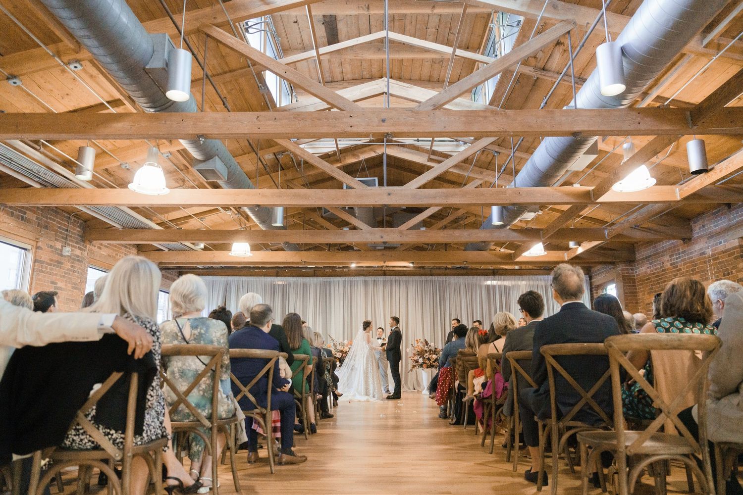 Guests seated in wooden chairs watch a wedding ceremony in a rustic brick venue with exposed beams and white draping.