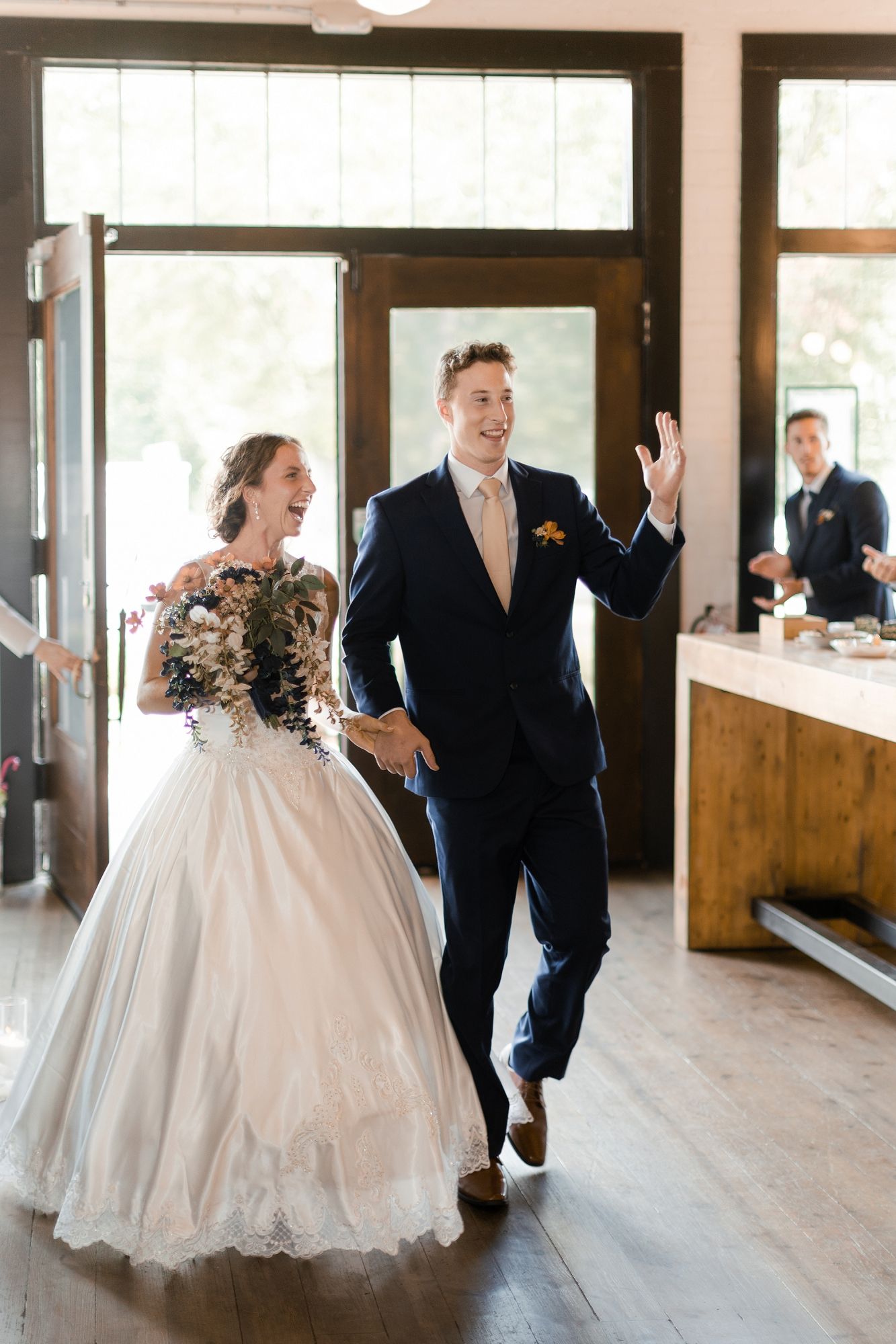 Joyful bride and groom in navy suit make grand entrance into rustic wedding reception venue.