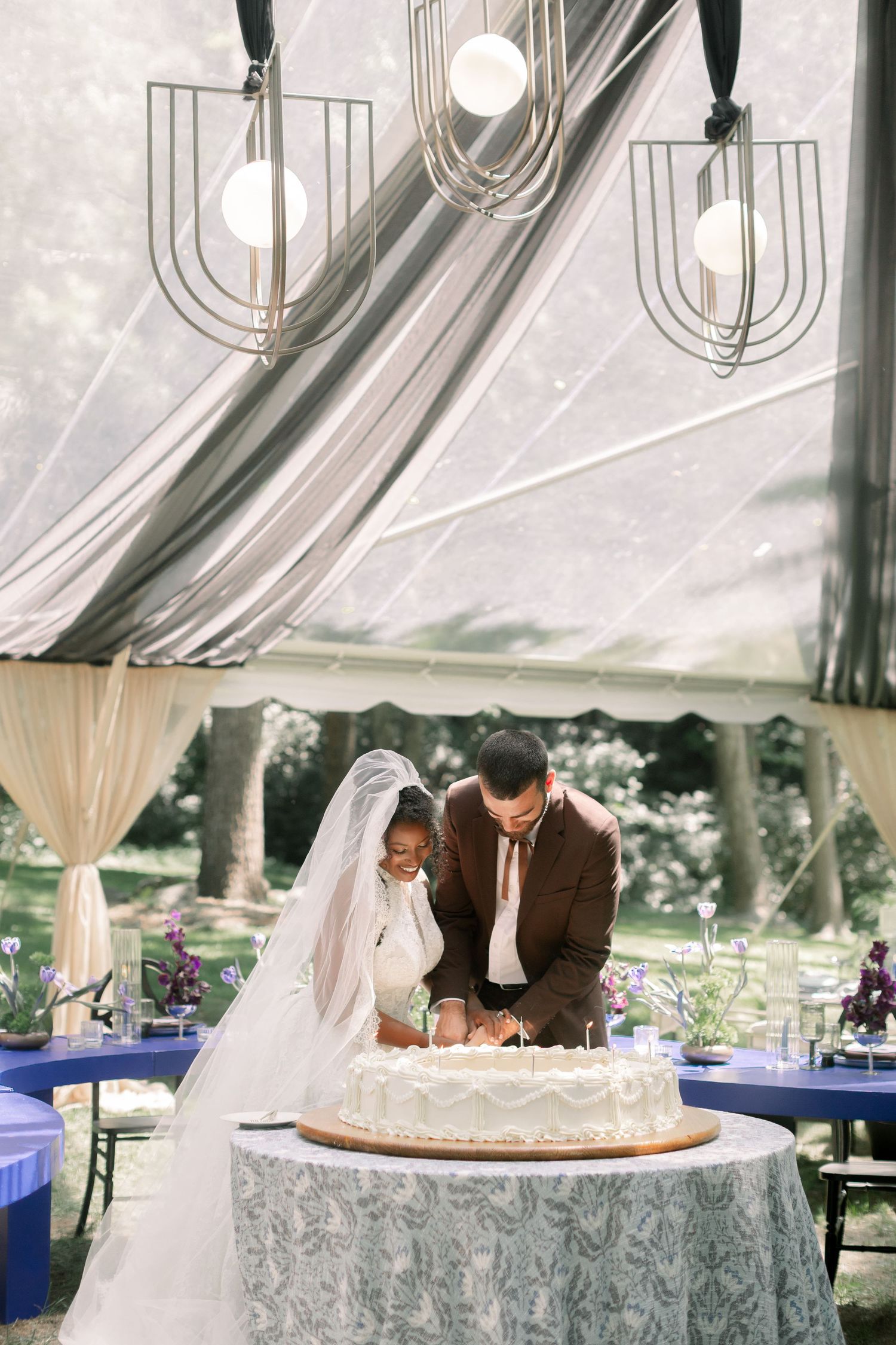 Bride and groom cutting white wedding cake under elegant tent with geometric pendant lights and blue reception tables.