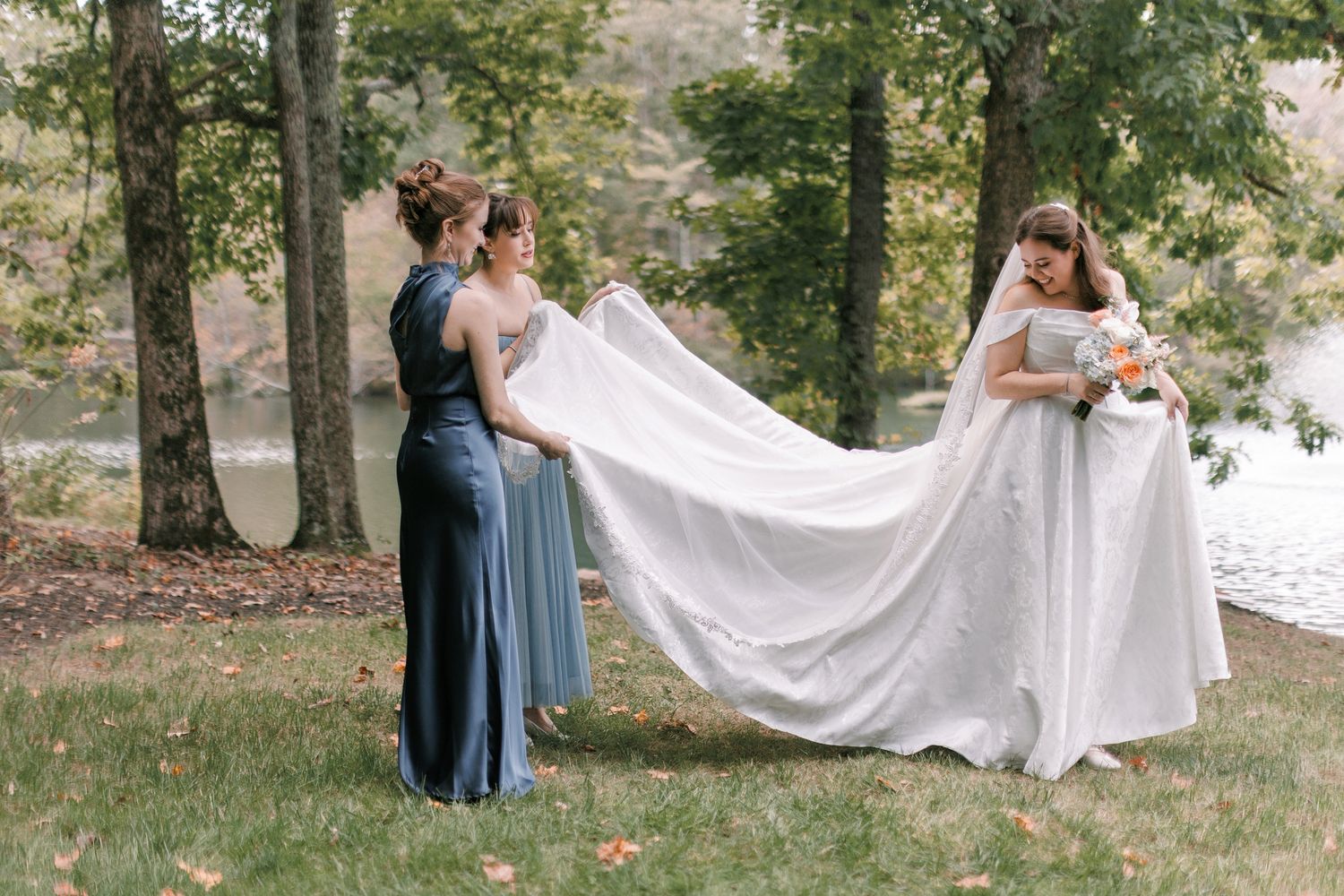 Bridesmaid in blue gown helps spread the bride's long white wedding veil outdoors near a serene lake.