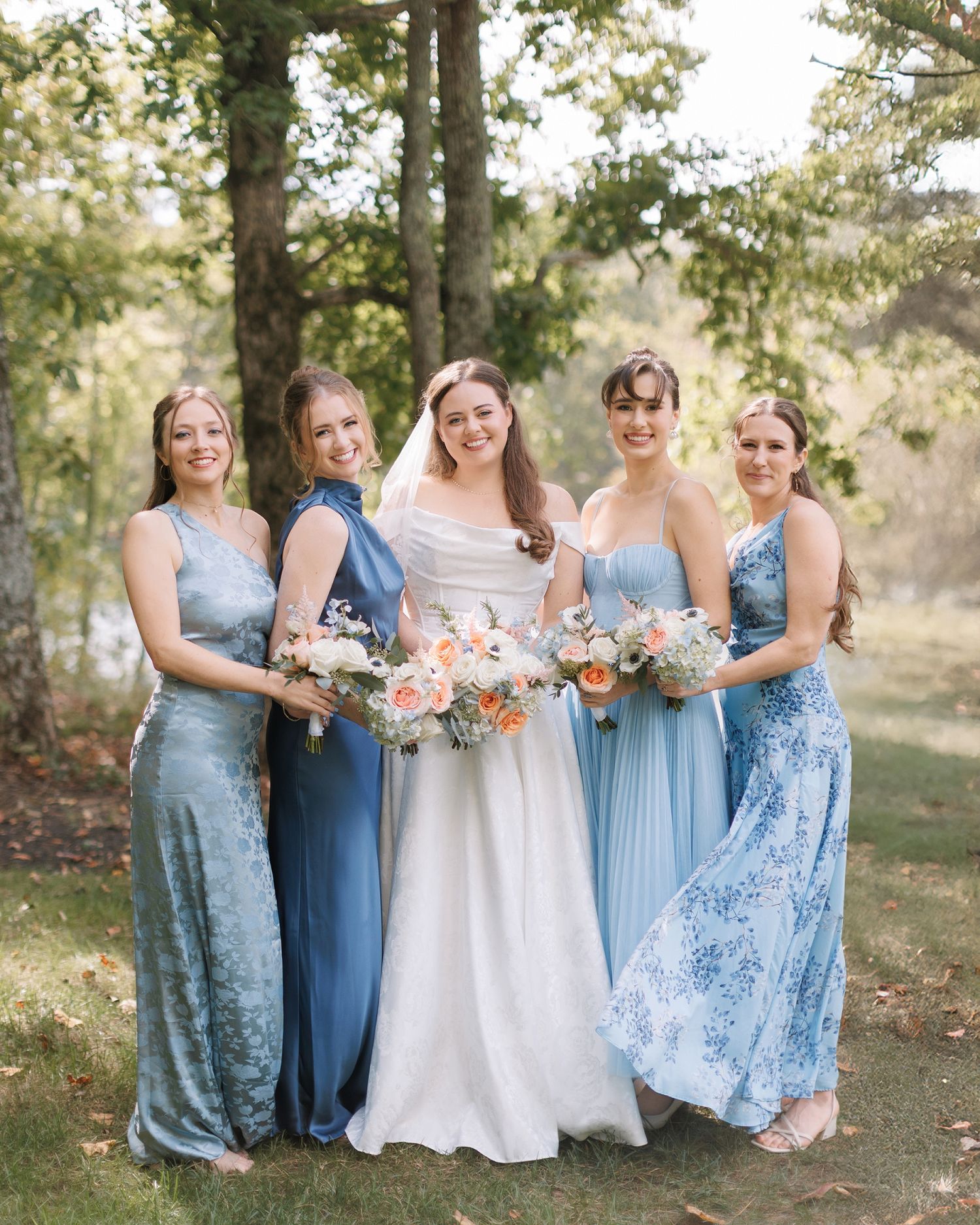 Bride in white gown poses with four bridesmaids in blue dresses holding floral bouquets outdoors.