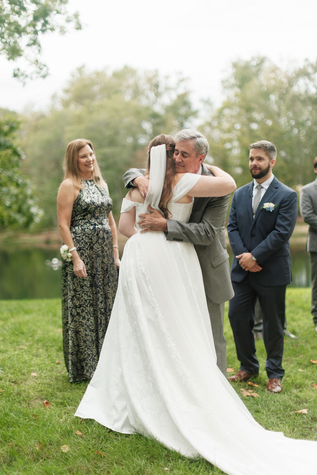 Father embraces bride in wedding gown as mother watches during outdoor wedding ceremony handoff.