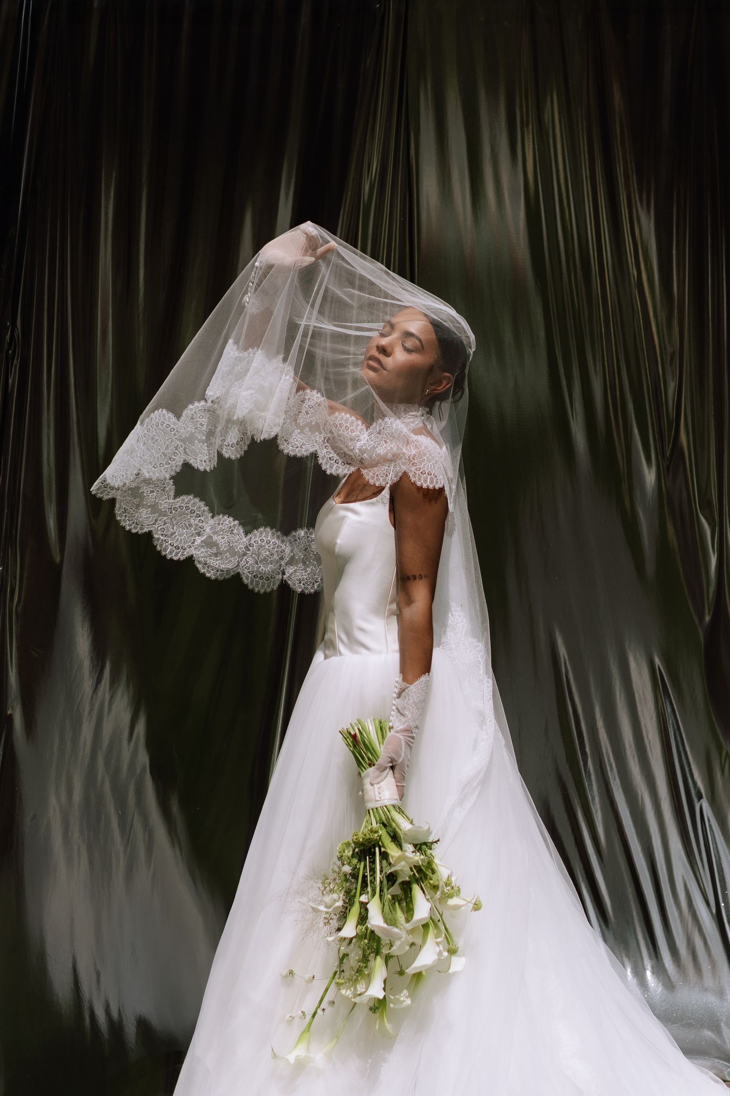 Bride in white gown and lace veil holds calla lily bouquet, posing gracefully against draped green backdrop.