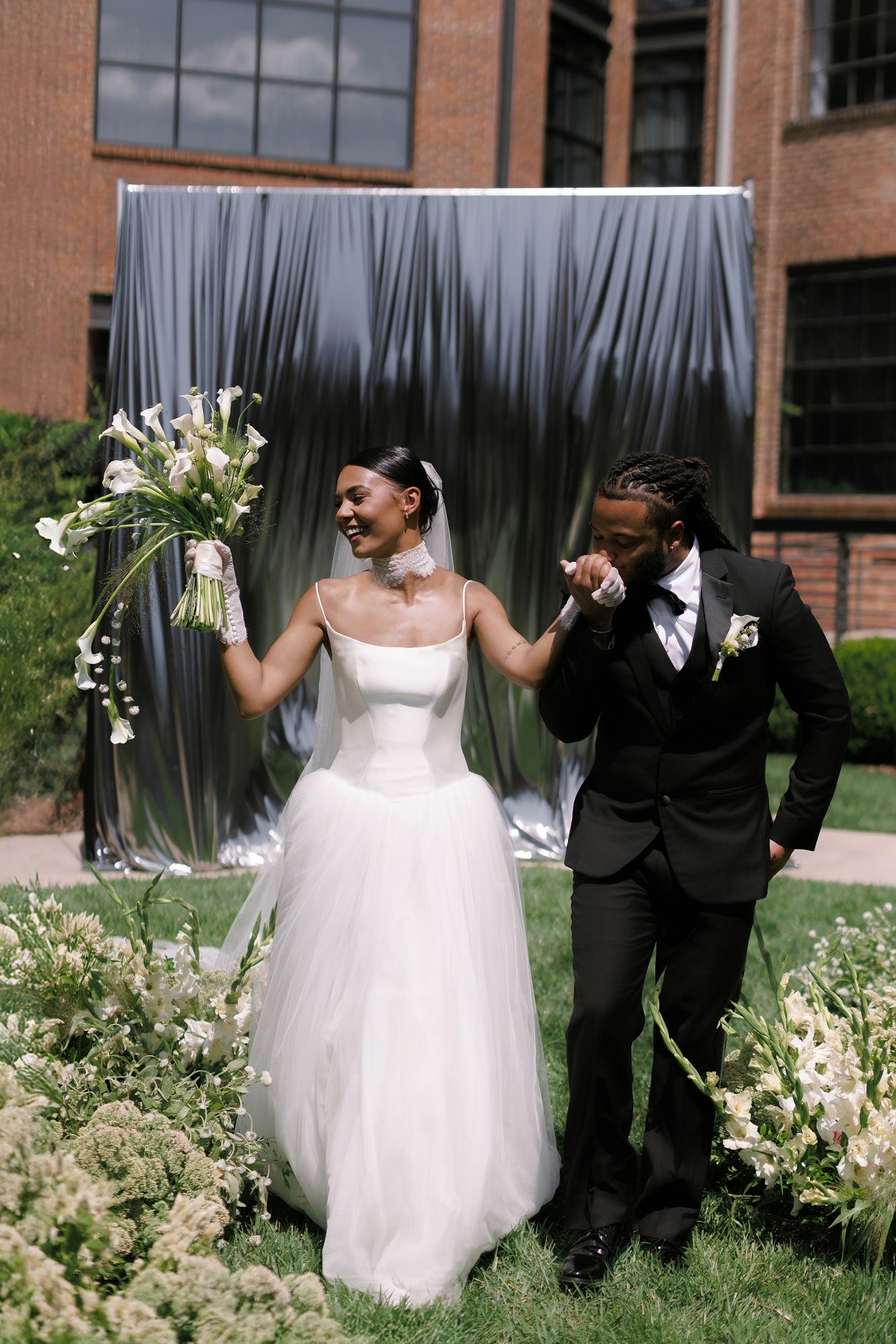 Bride and groom stand before a silver draped backdrop outdoors, she holds white calla lilies as he kisses her hand.
