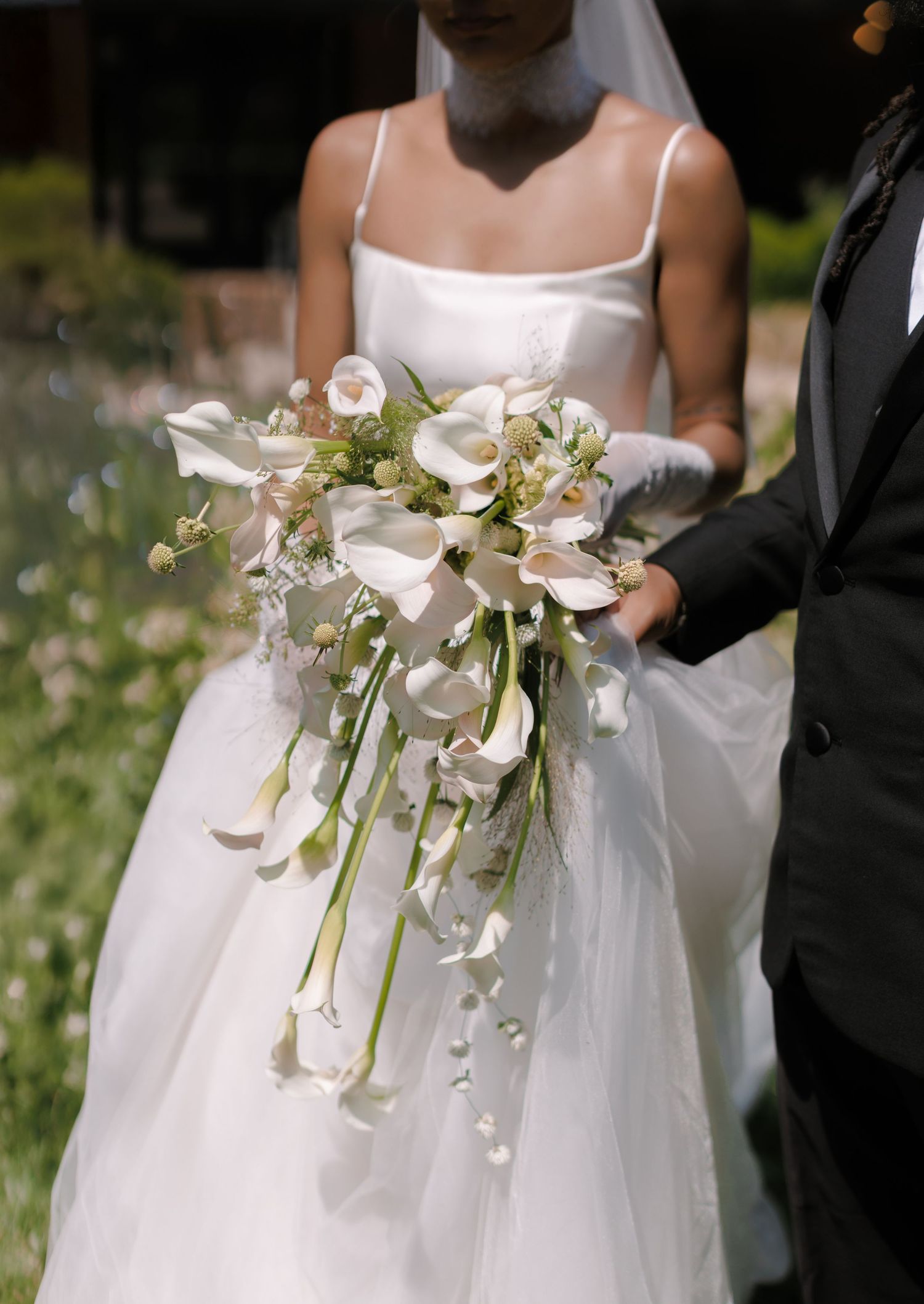 Bride in white gown holding elegant cascading calla lily bouquet with greenery beside groom in black tuxedo.