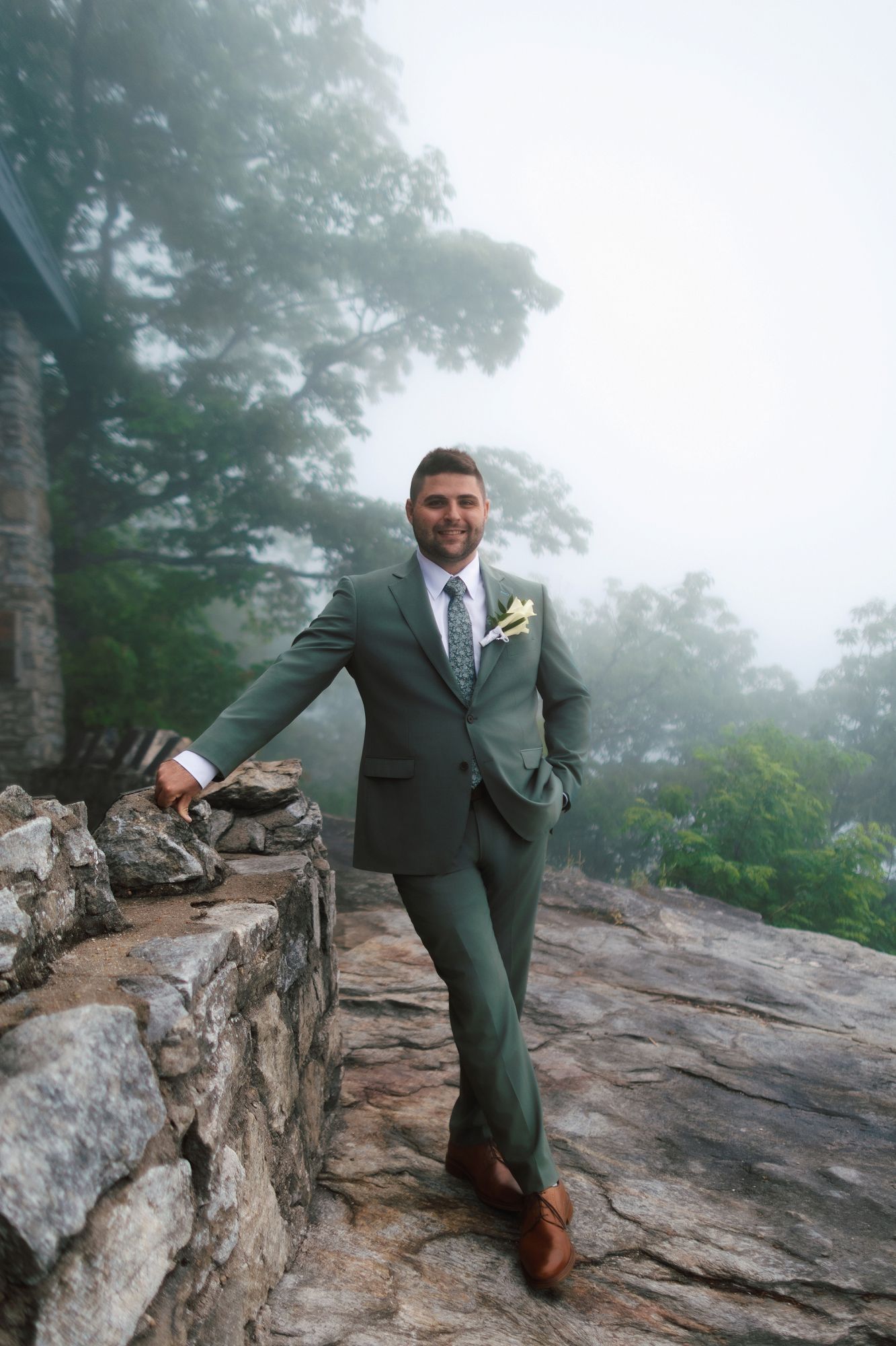 Smiling groom in green suit with boutonniere posing on rocky overlook surrounded by misty trees.