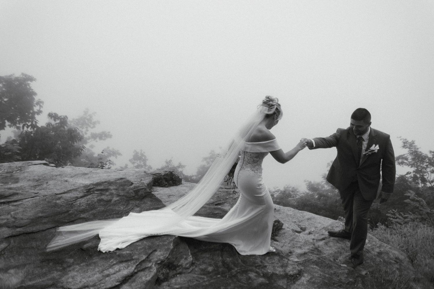 Black and white photo of groom helping bride navigate rocky terrain in foggy mountain landscape after wedding.