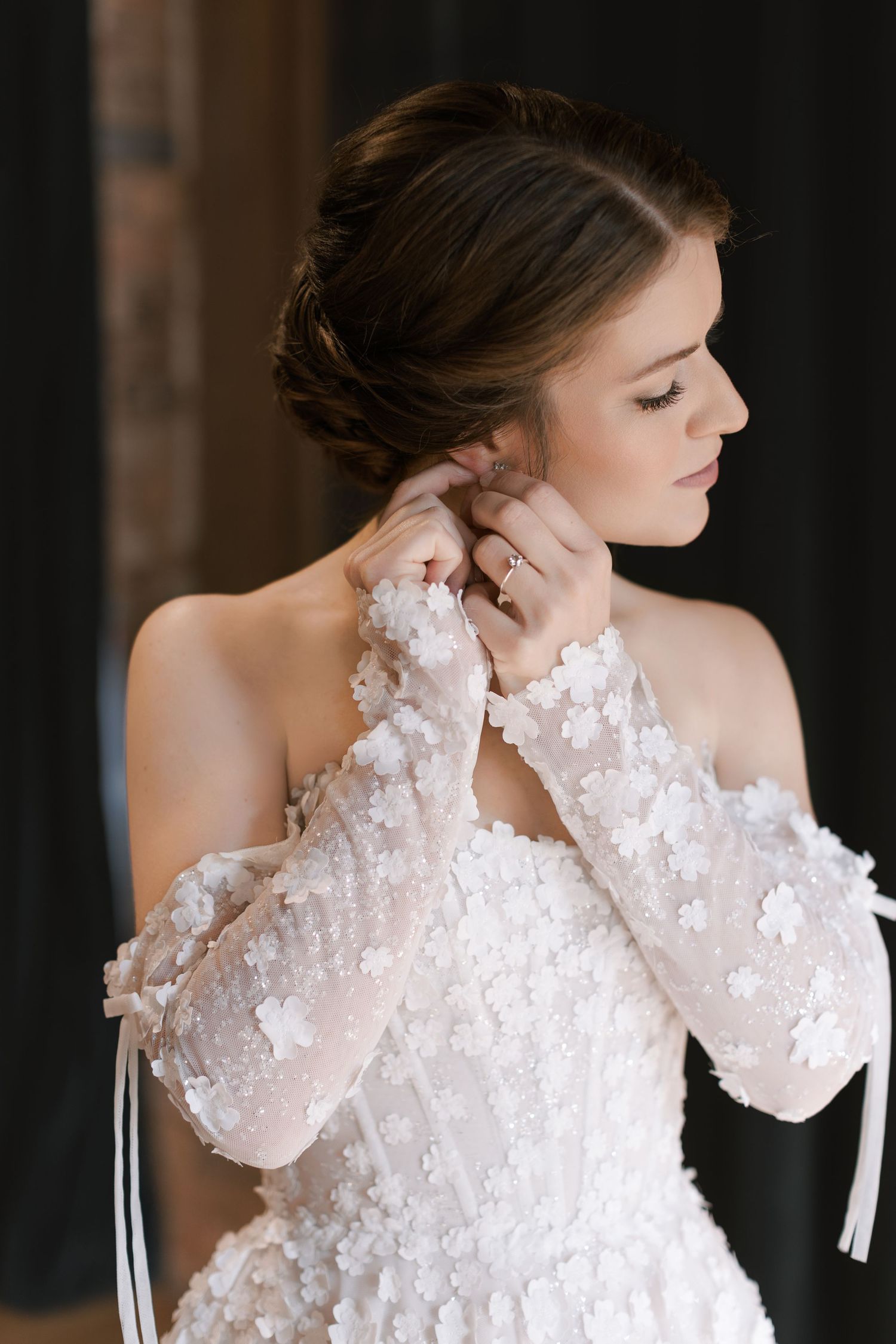 Bride in floral lace off-shoulder gown adjusting earring, elegant updo hairstyle, soft bridal portrait.