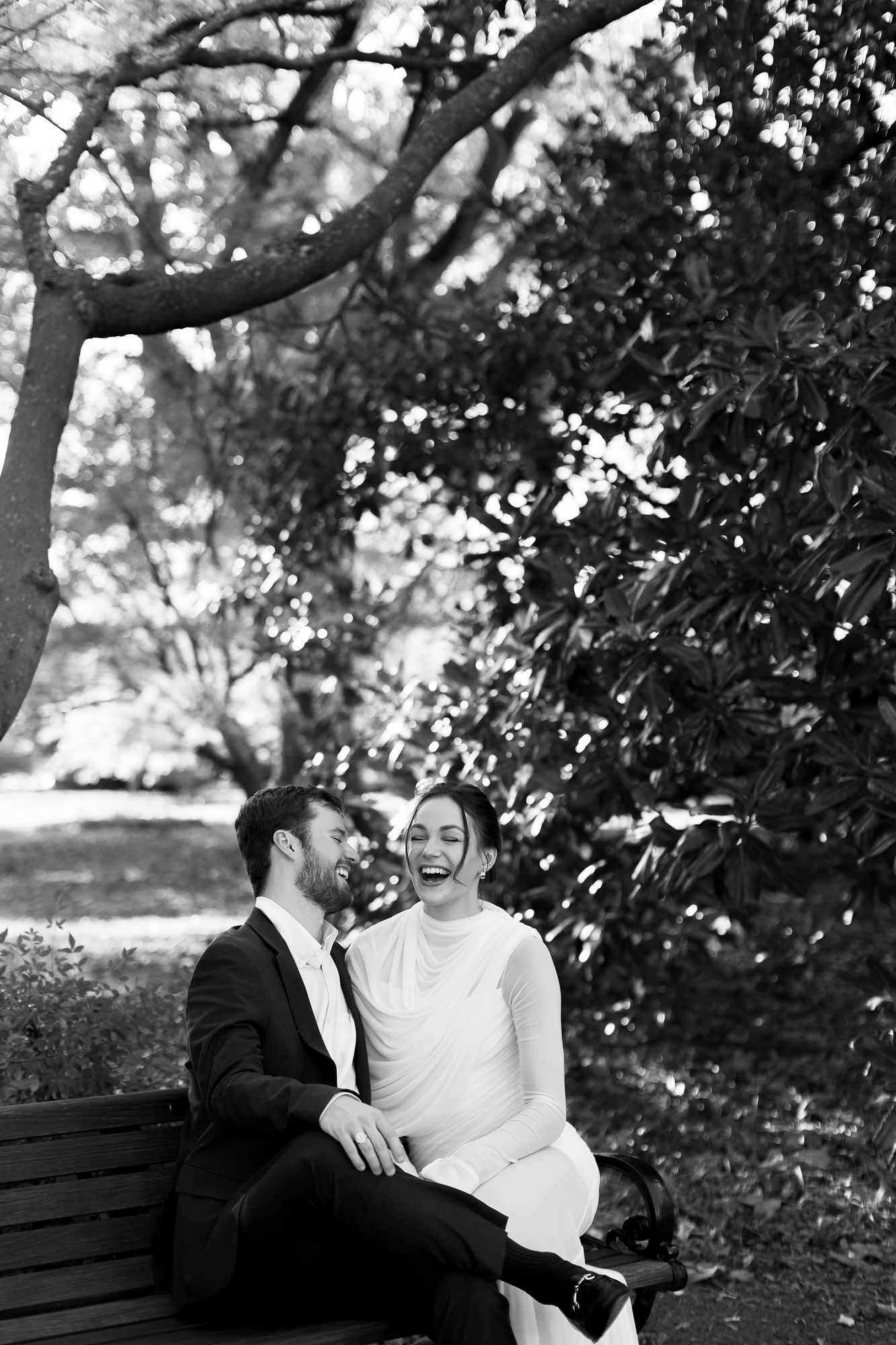 A romantic couple sits together on a park bench surrounded by trees in a black and white photo series.