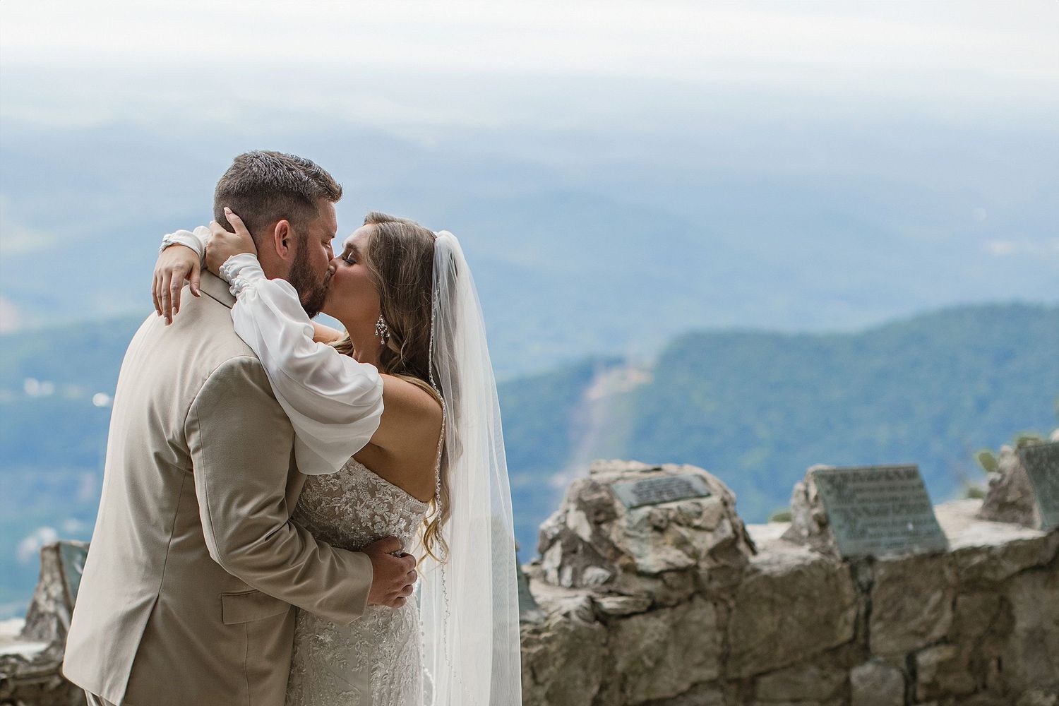 Wedding couple embraces against a scenic mountain backdrop with stone wall details.