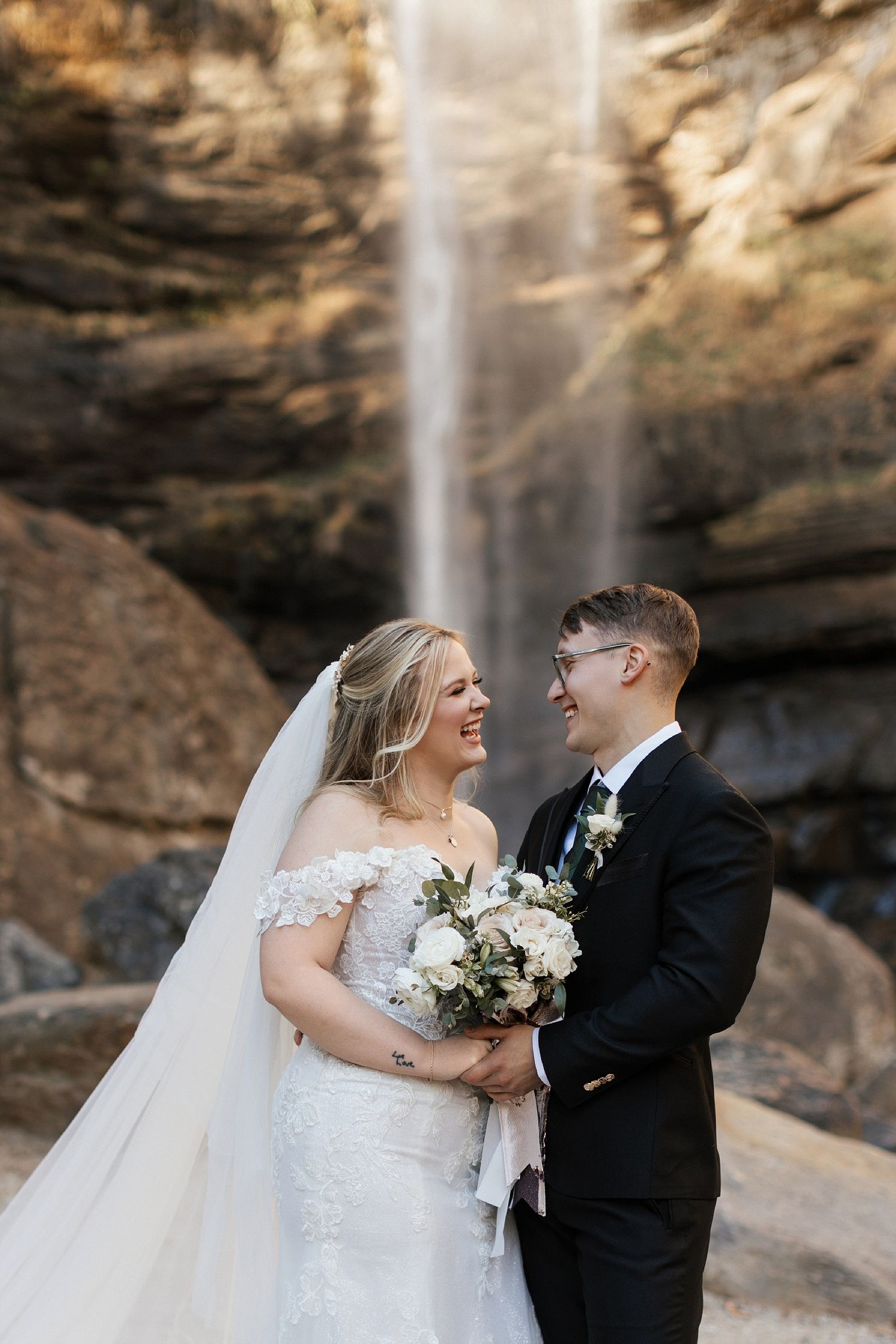 A couple in wedding attire shares a joyful moment in front of a majestic waterfall backdrop.