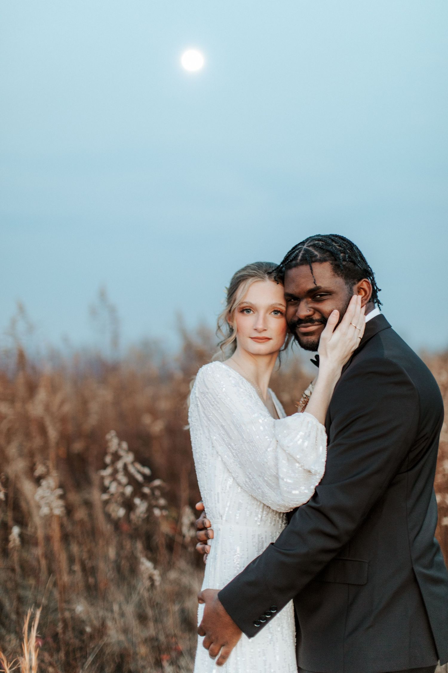 A couple embraces in front of a dreamy sunlit field with dried tall grass and pale blue sky.