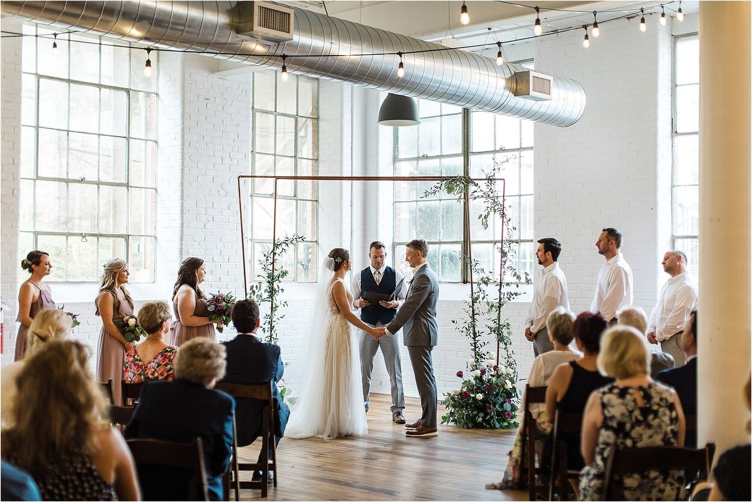 Modern indoor wedding ceremony with exposed ductwork and large windows in an industrial-style venue.