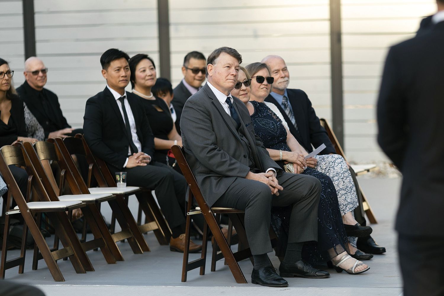 Wedding guests in formal attire seated on wooden folding chairs during an outdoor ceremony.