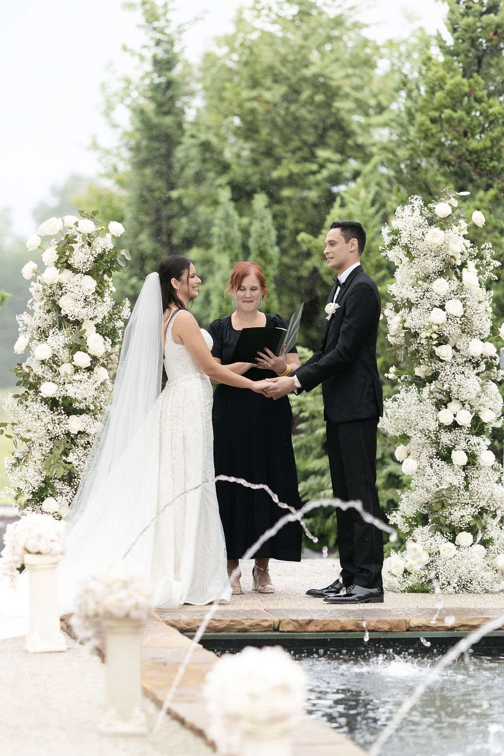 Wedding ceremony setup with white floral arches and fountain in garden setting.