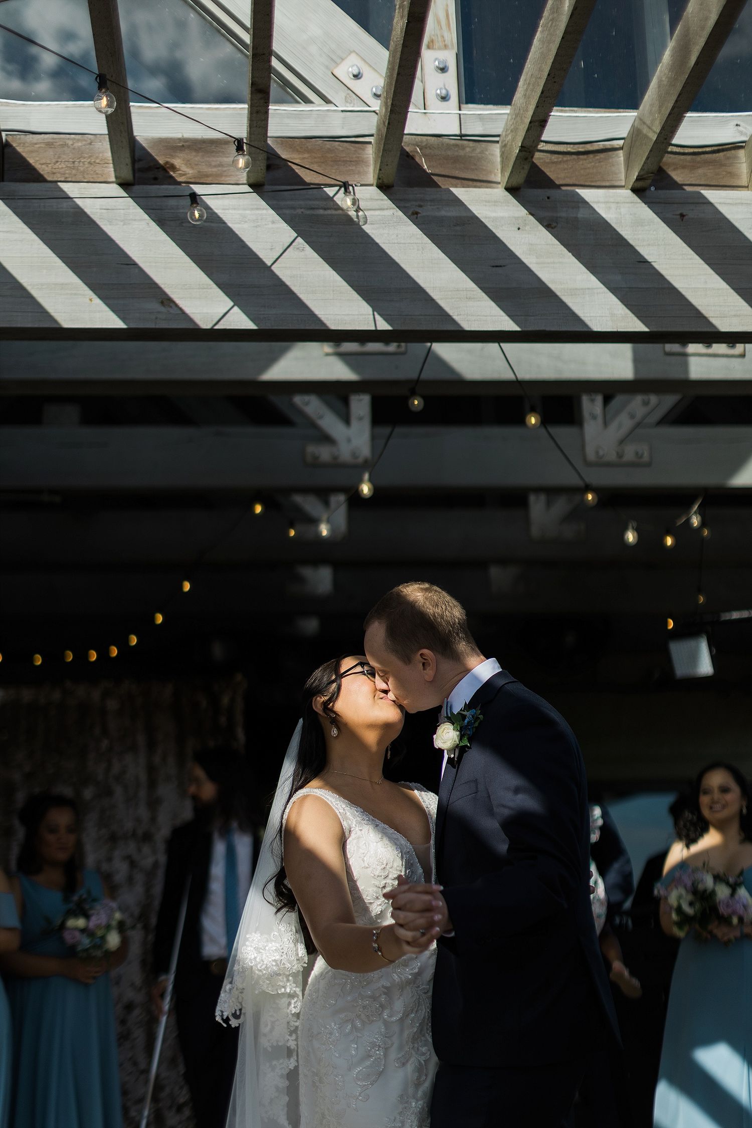 A romantic wedding moment under a wooden pergola with dramatic shadows and string lights.