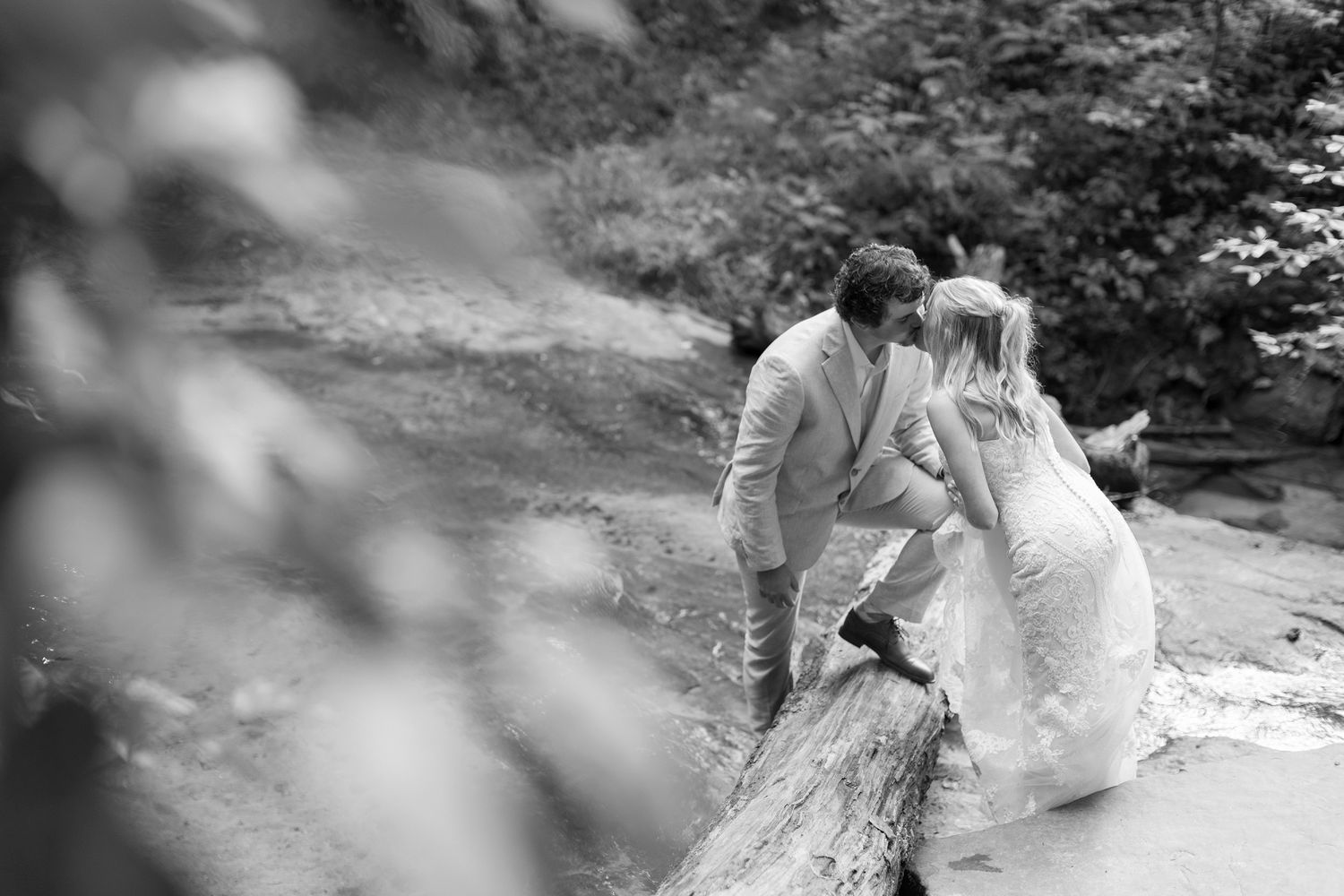 A black and white romantic wedding photo captures a couple leaning on a stone bridge in a dreamy forest setting.