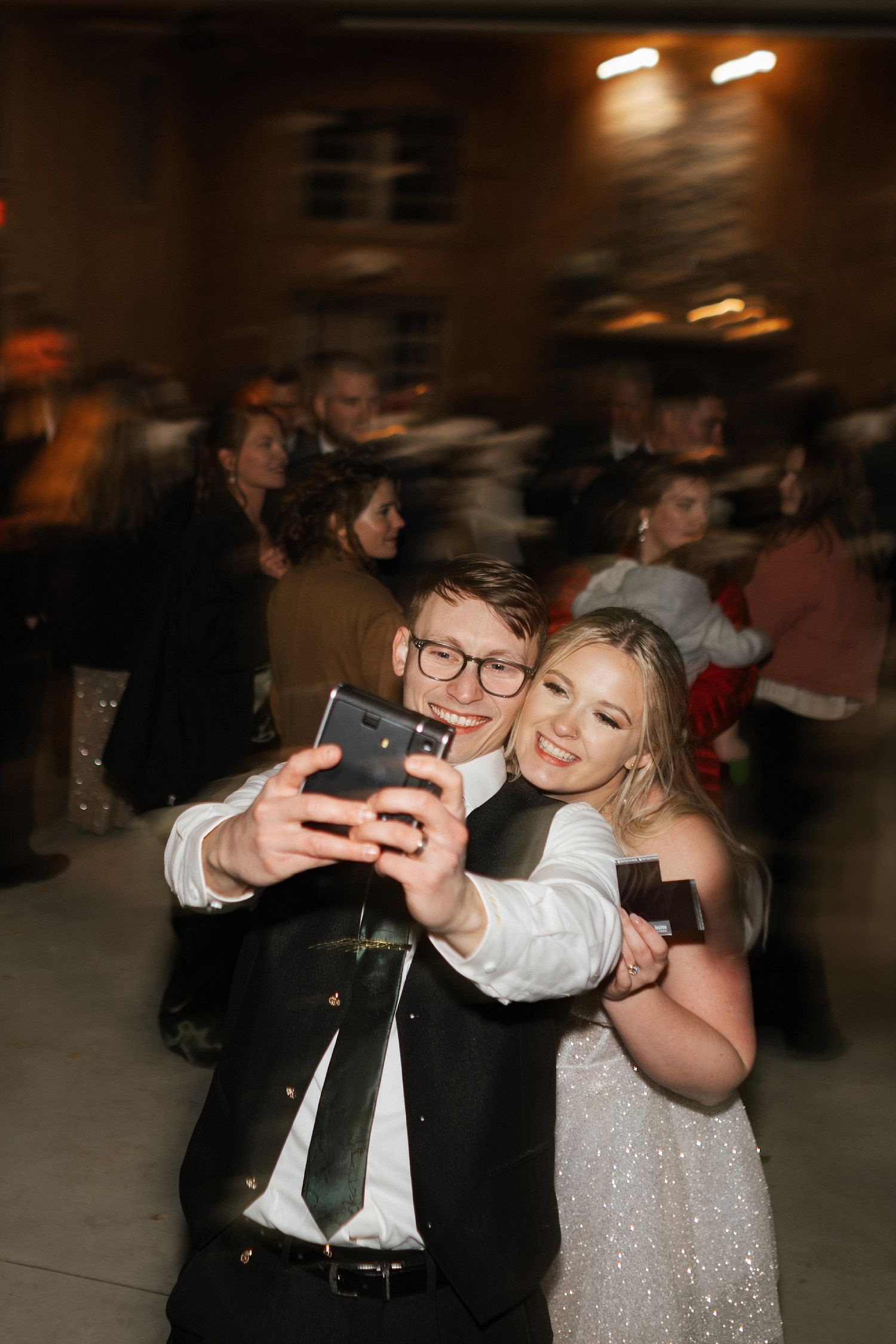 A pair in formal attire takes a selfie together at a dimly lit party venue with sparkly decorations.