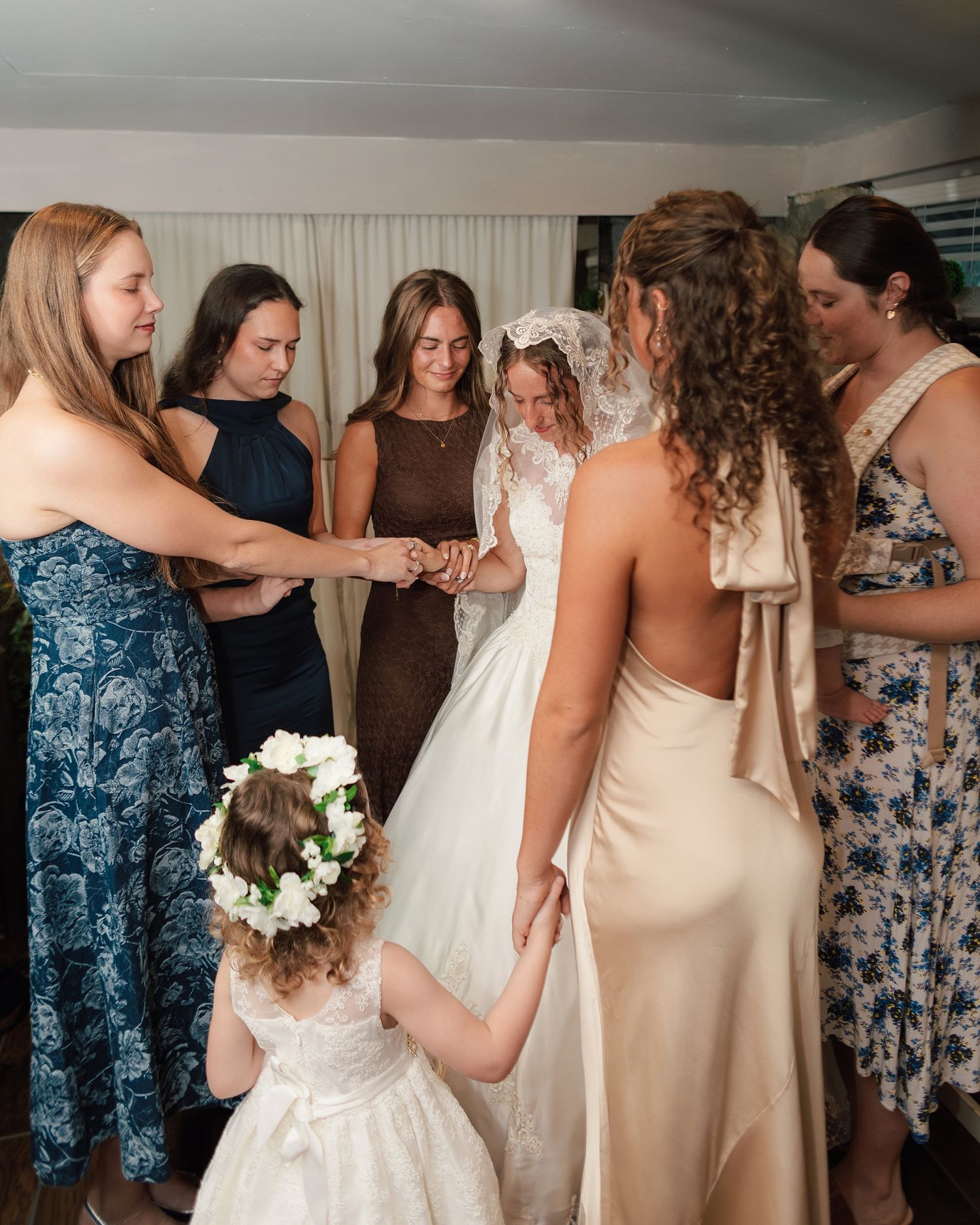 Bride in white lace gown and veil surrounded by bridesmaids and flower girl holding hands in a circle.