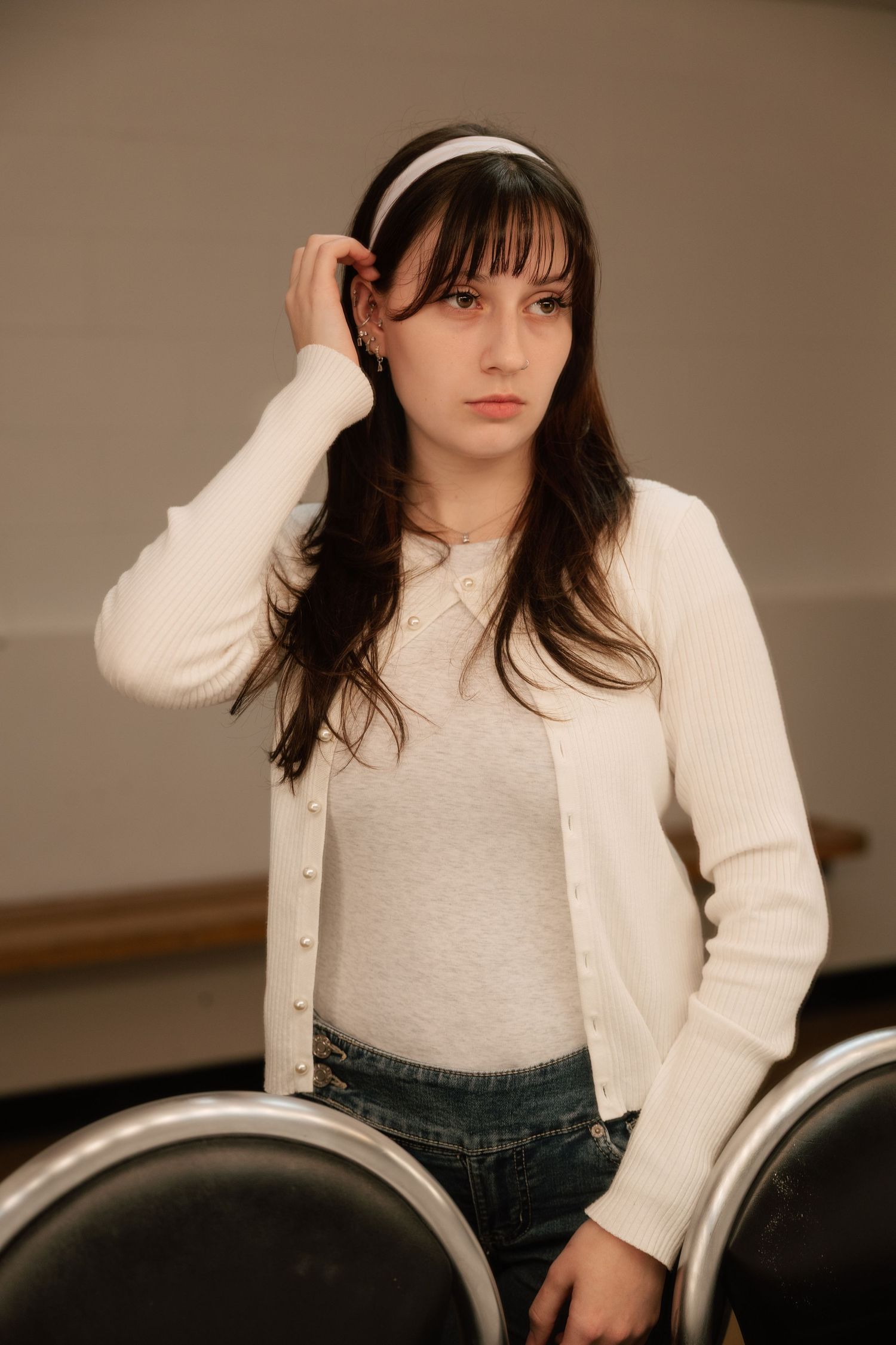 Young woman with dark hair and headband in white cardigan and jeans, posing thoughtfully near chairs.