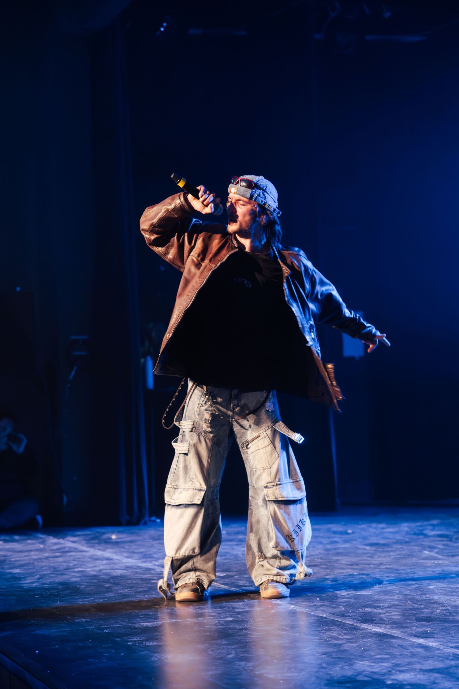 Performer singing into microphone on stage wearing oversized brown jacket and cargo pants under blue stage lighting.