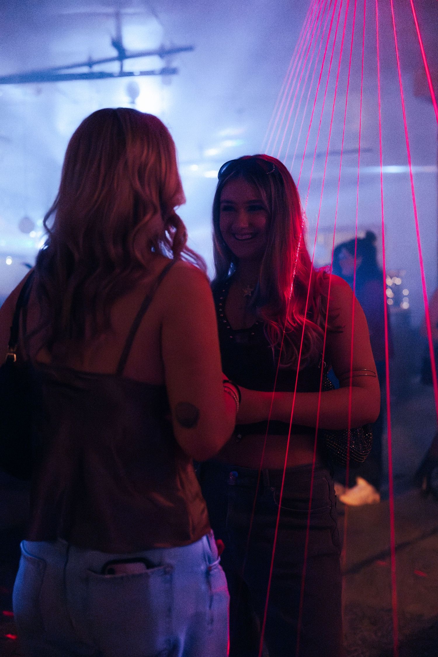 Two women chat and smile amid vibrant red laser lights at a nightclub event.