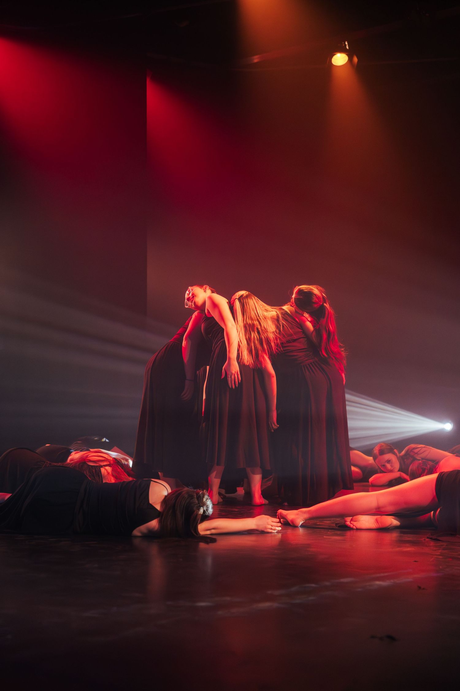 Dramatic dance performance with dancers lying on floor while others stand in dark gowns under red spotlight beams.