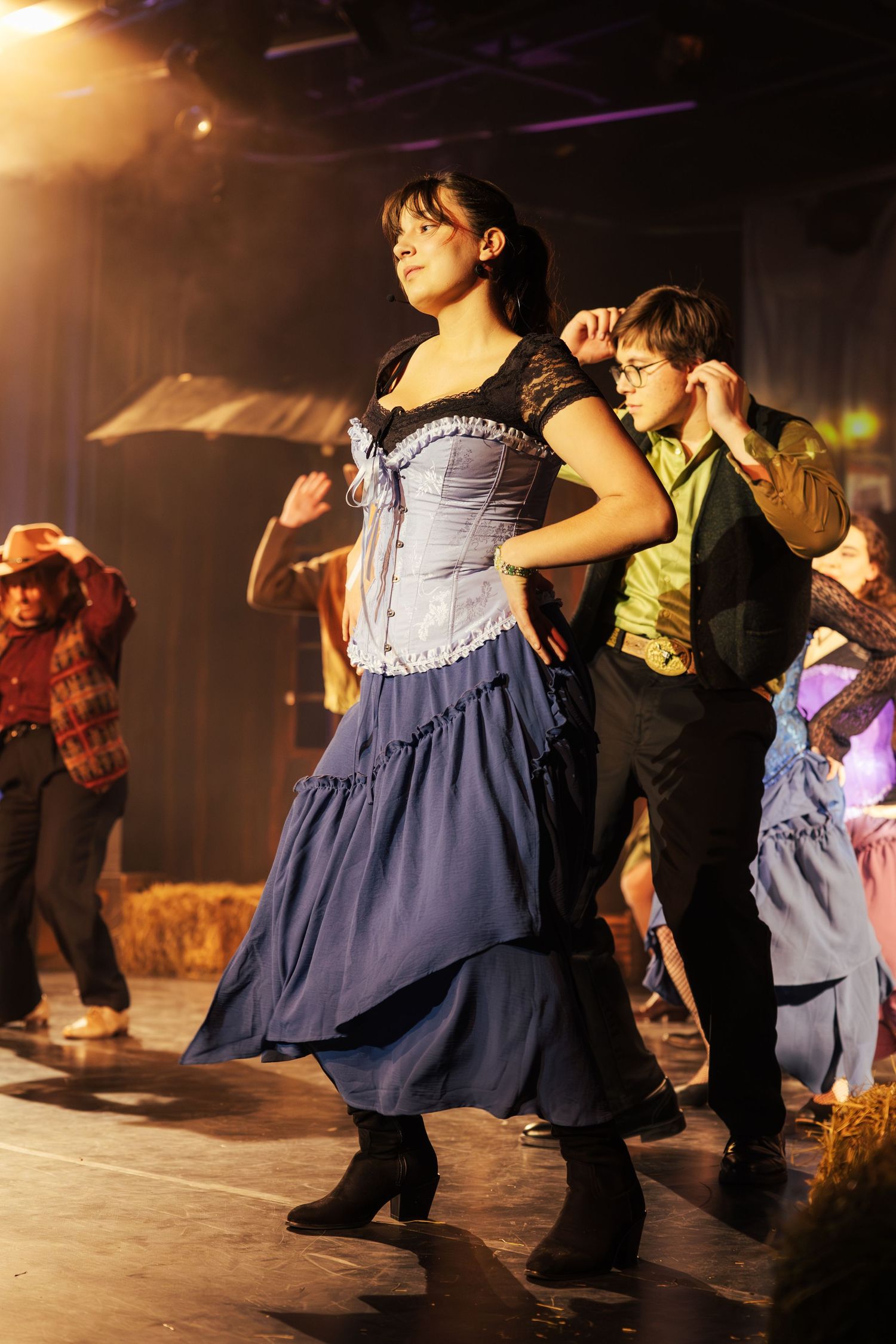 A woman in a blue corset and flowing skirt leads a group of dancers in a western-themed stage performance.