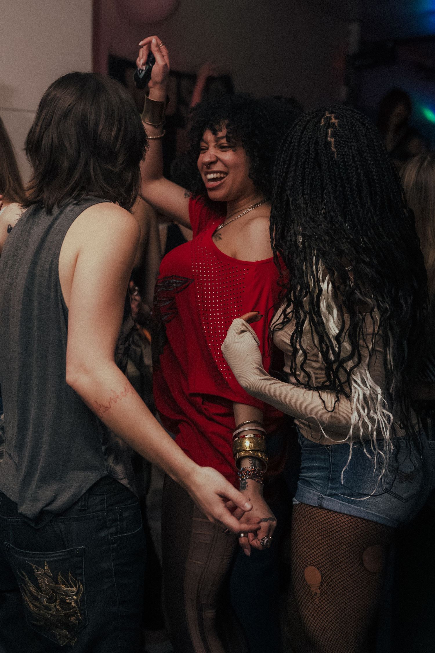 Three people laughing and dancing together at a party, woman in red top at center.