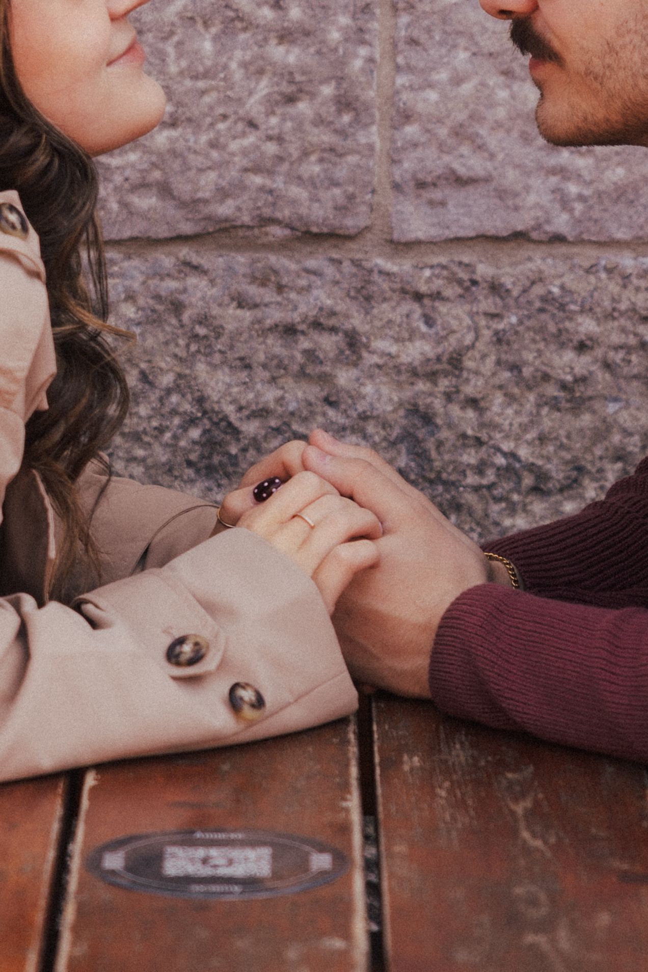A couple holds hands across a wooden table, leaning in close for a kiss outdoors.